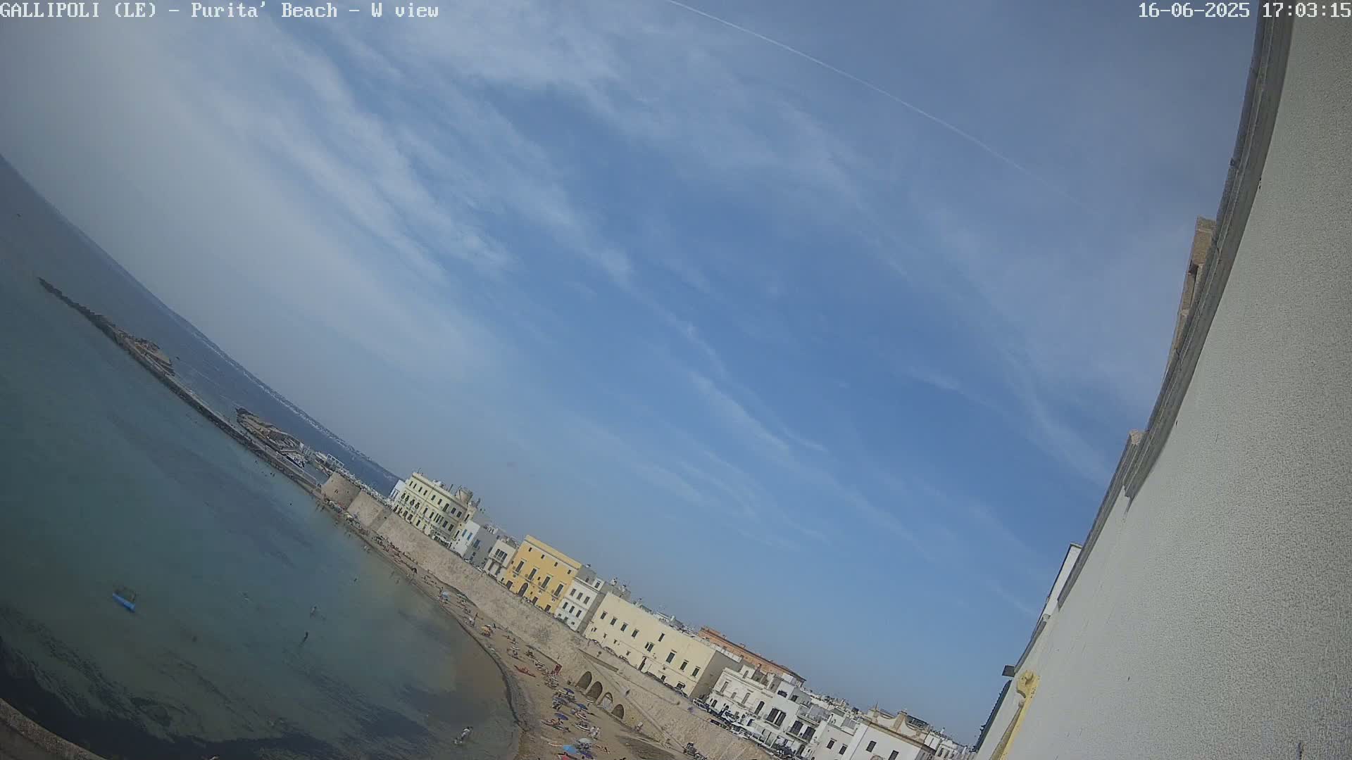 A mostly sunny day shows a beach with light-colored buildings along the shoreline and a calm, clear sea.