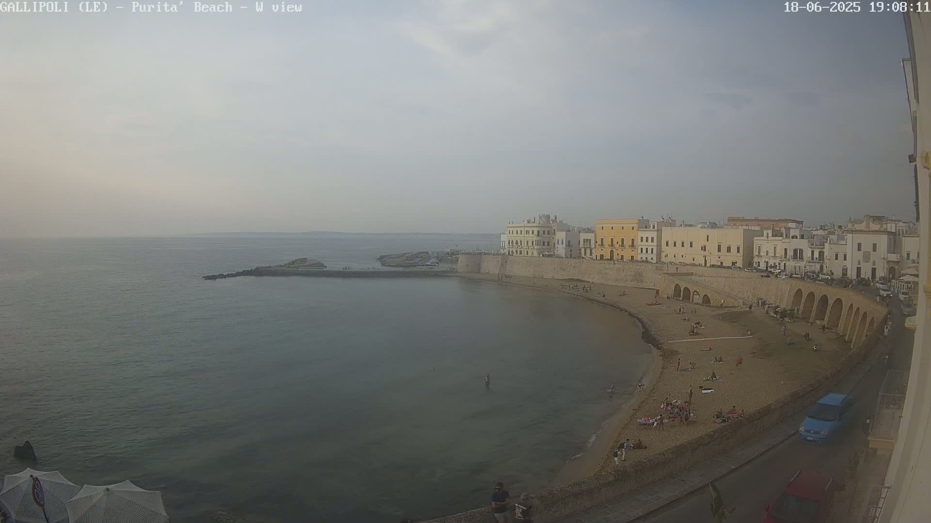 A hazy, overcast view of a sandy beach curving around a calm sea, backed by a line of buildings along a stone wall with arched structures.