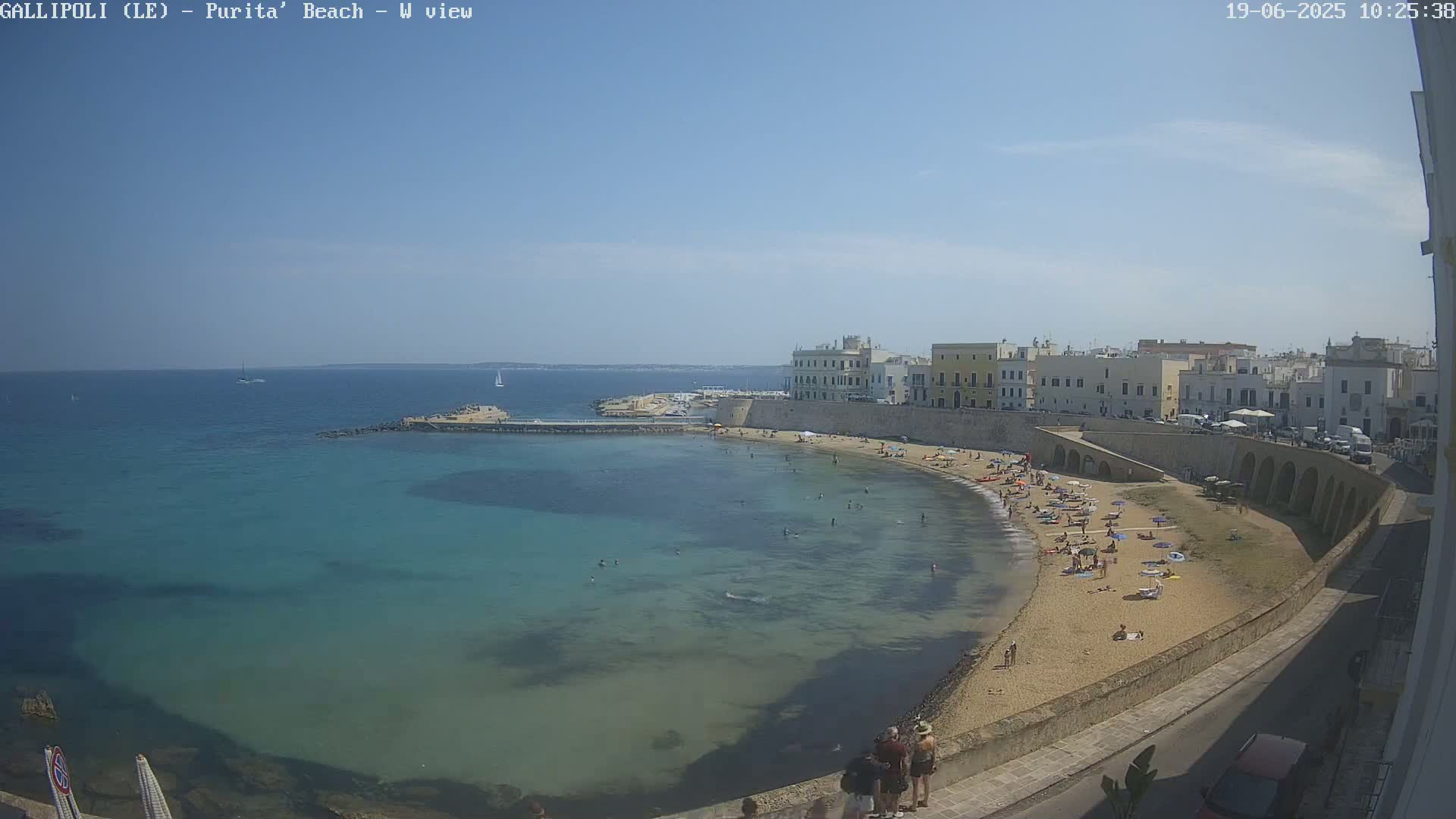 A sandy beach with many people on it curves around a calm, clear teal sea next to a town of light-colored buildings under a sunny sky.