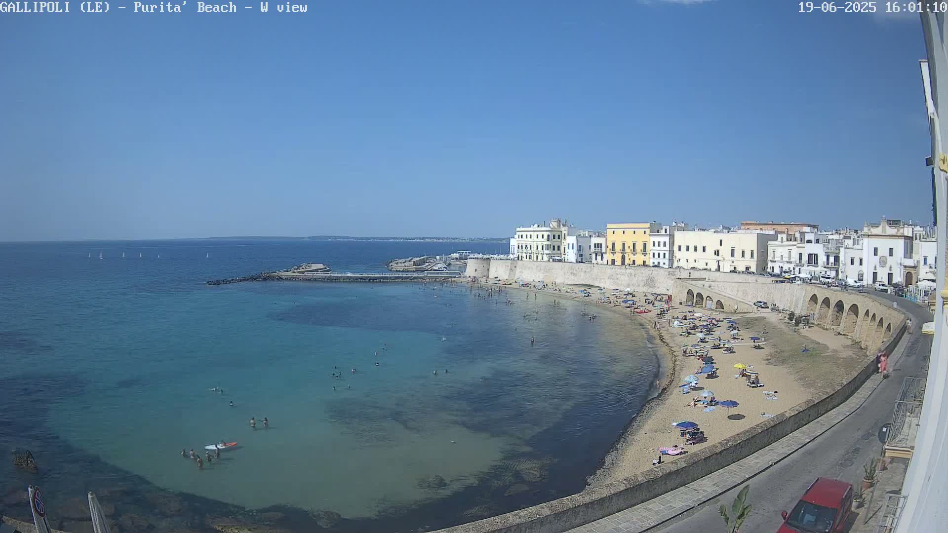 A clear, sunny day shows a beach with many people, calm blue-green water, and a seaside town with light-colored buildings and arched walls.