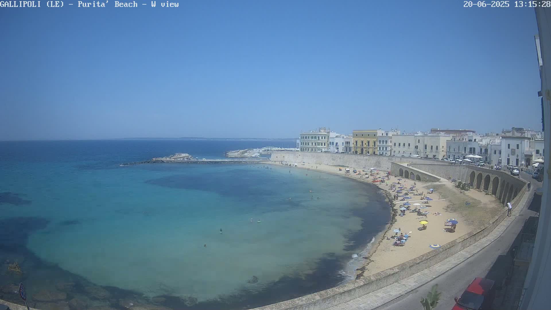 A sandy beach curves around a calm, turquoise bay, lined with buildings and people under a clear blue sky.