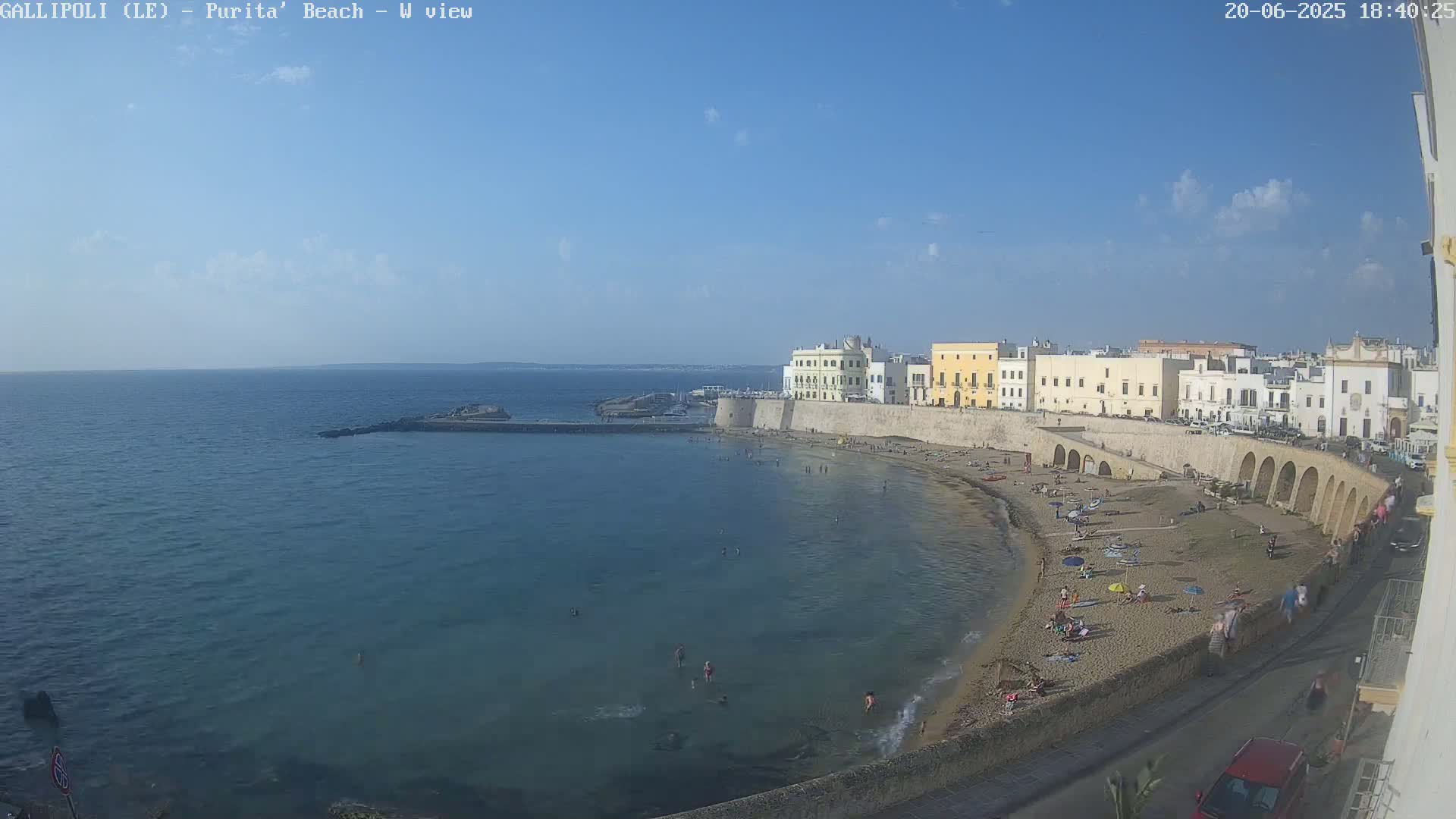 A sandy beach with bathers is nestled beside a calm, blue sea and a walled city under a clear, bright sky.