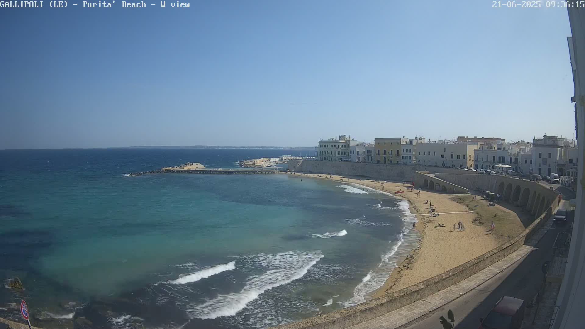 A sandy beach with light wave action curves around a calm, turquoise sea next to a walled town under a clear, sunny sky.