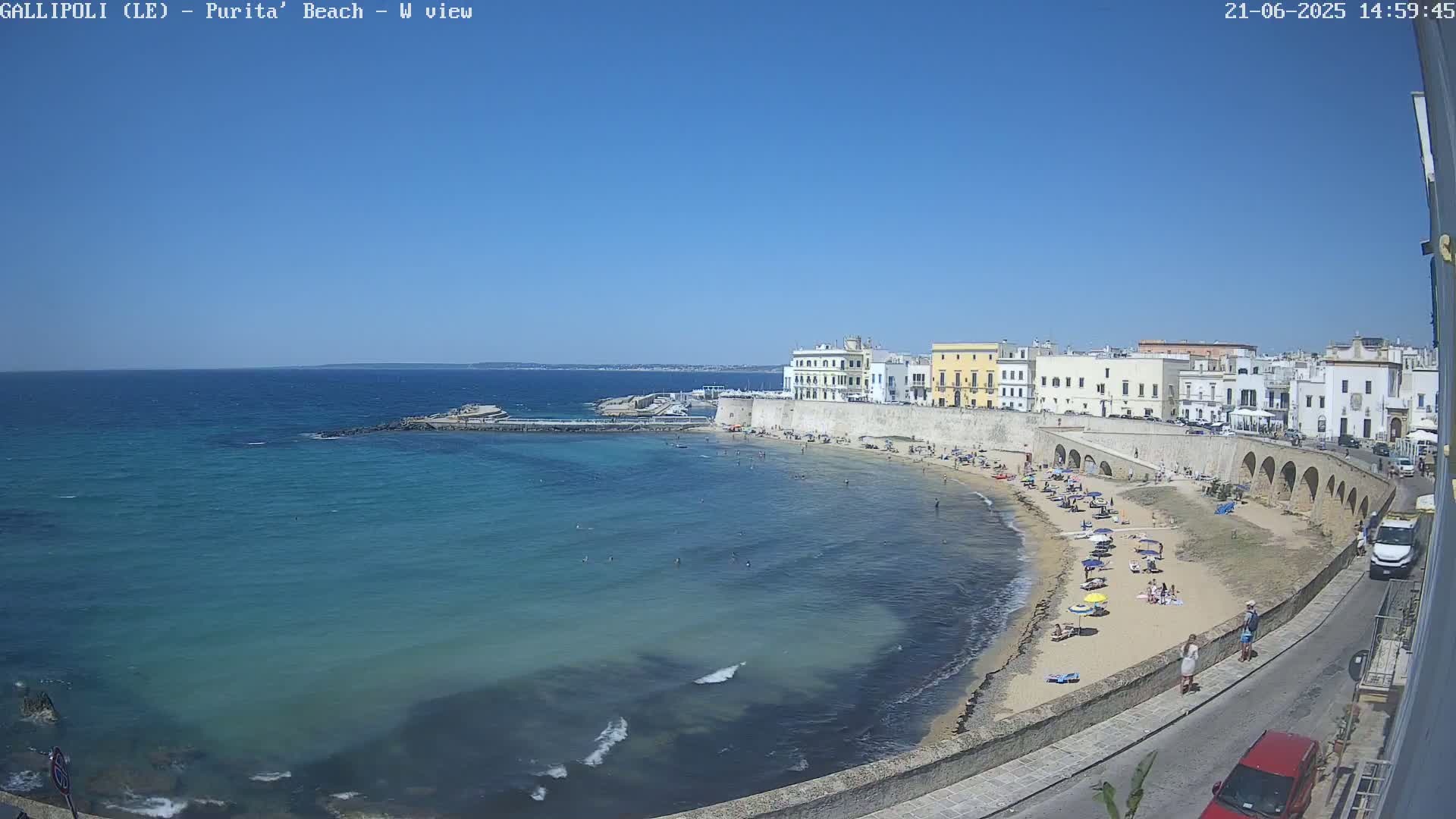 A sandy beach with sunbathers is situated below a waterfront town under a clear blue sky.