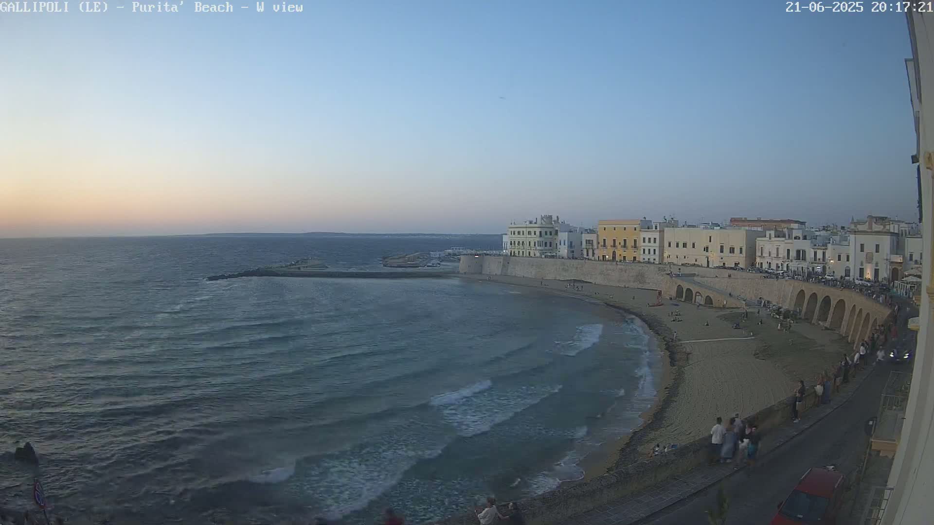 A sandy beach with gentle waves laps against a seawall bordering a town at twilight.