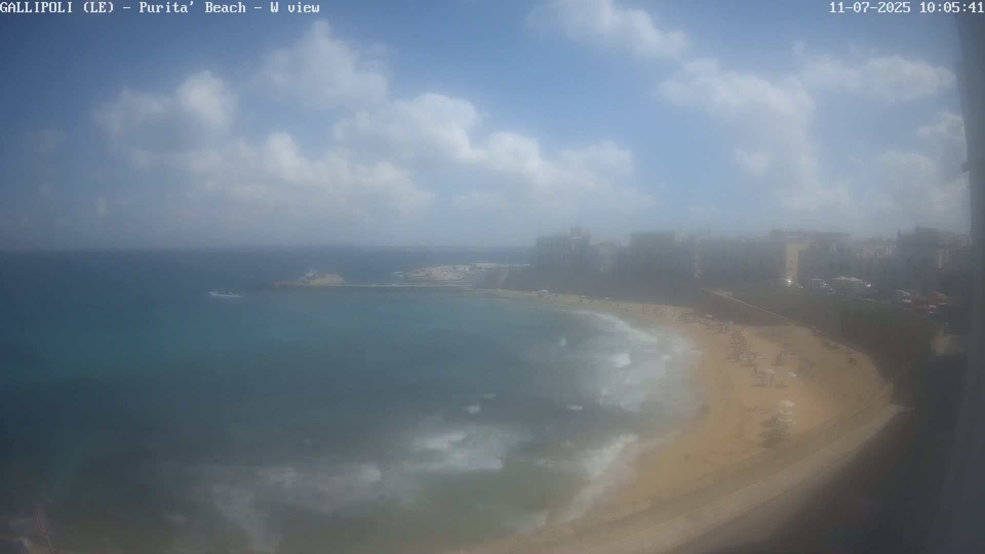 A partly cloudy day at a beach with light waves lapping a sandy shore, overlooking buildings in the distance.