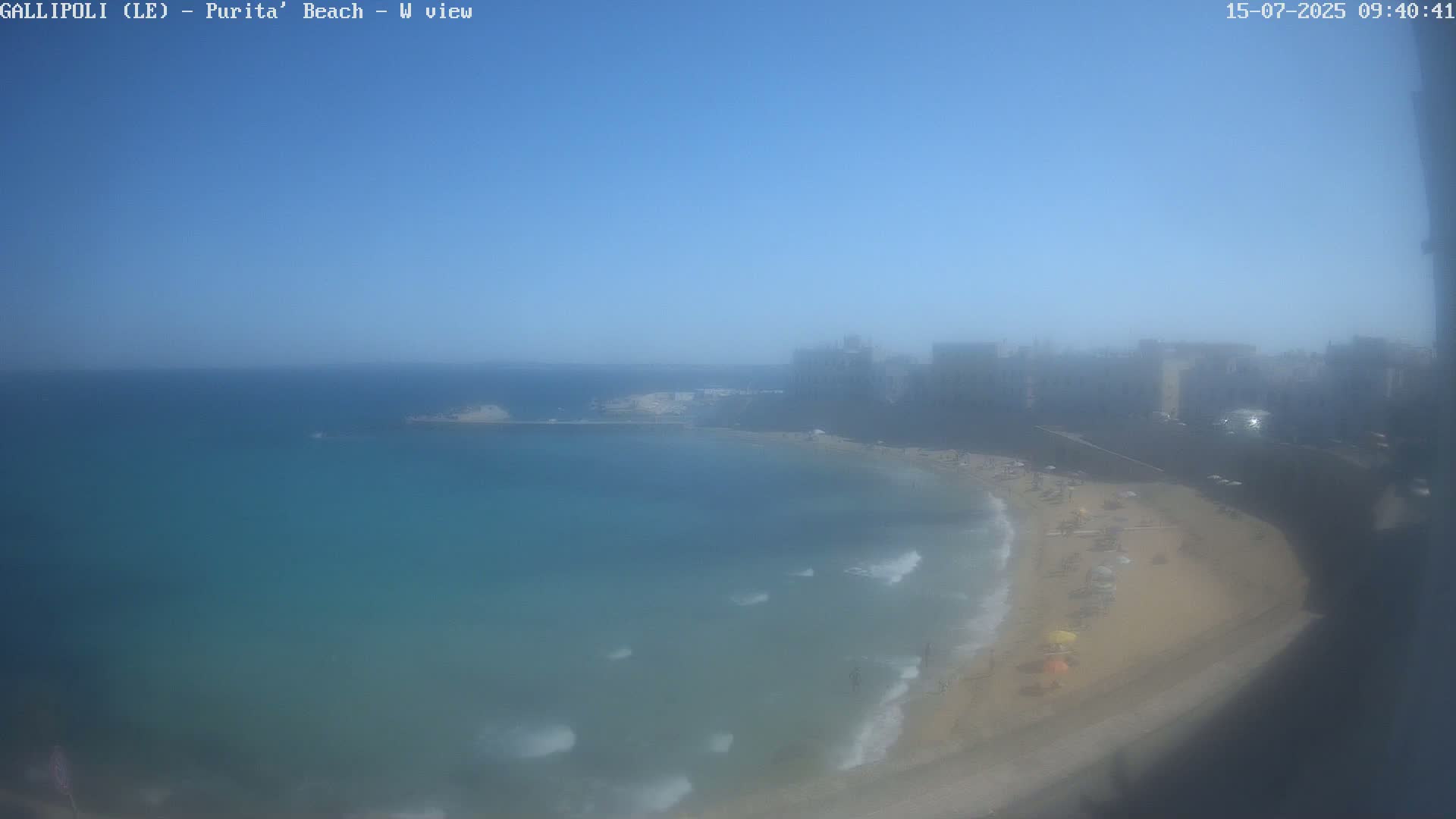 A hazy, sunny day shows a sandy beach with people and umbrellas, gentle waves lapping the shore, and buildings visible in the distance.
