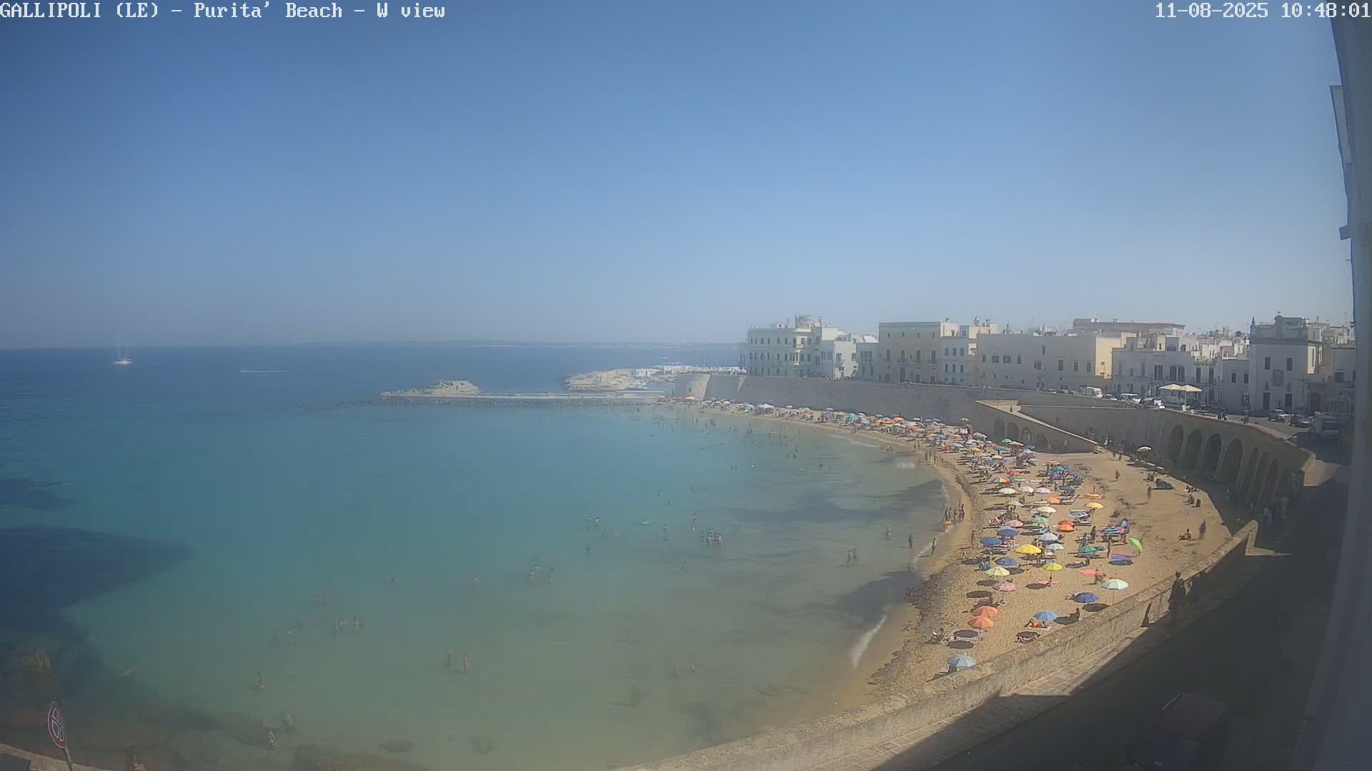 A sunny day at a crowded beach with clear turquoise water, lined with colorful umbrellas and bordered by a stone wall and buildings.