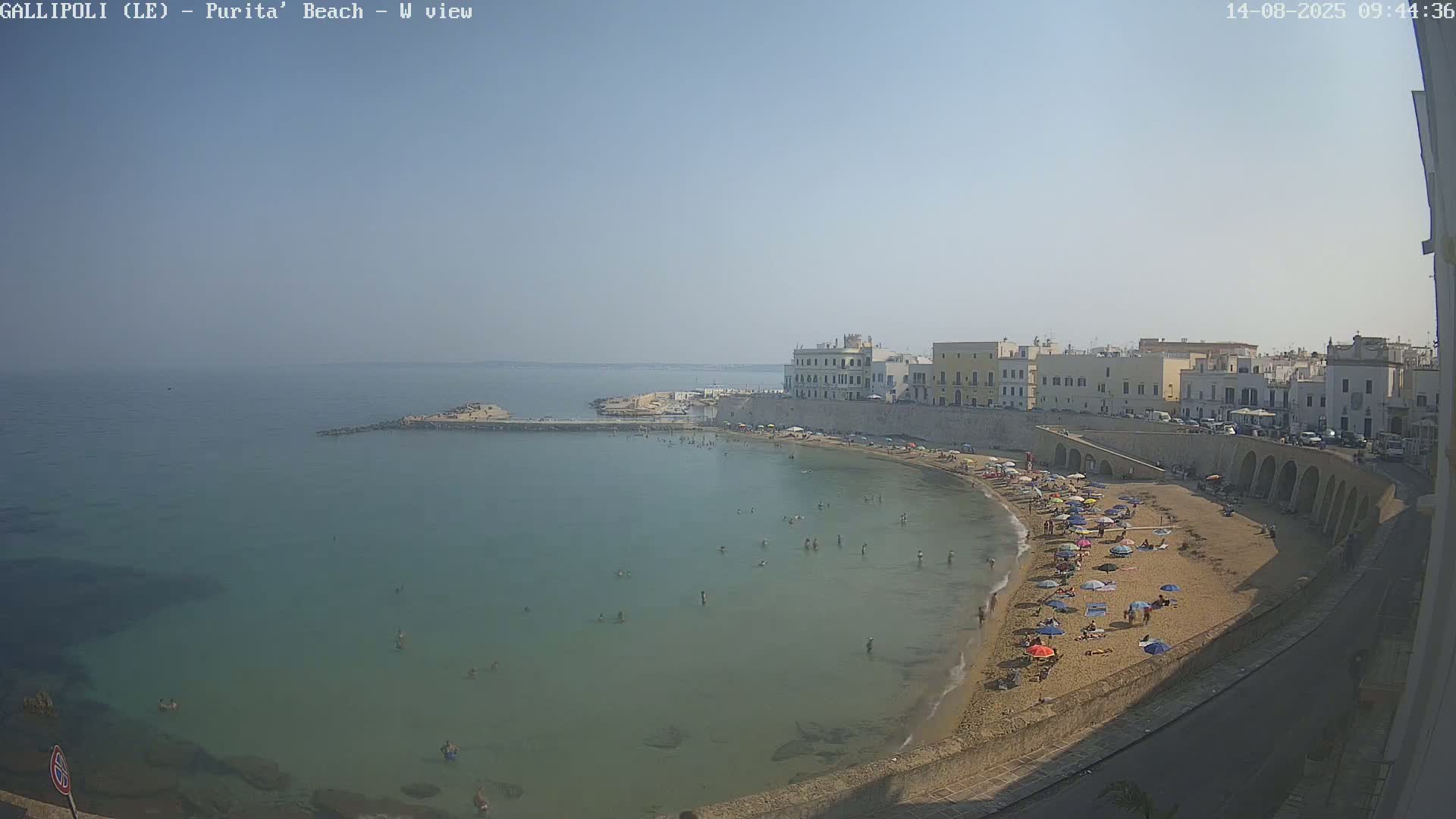 A sunny day at a sandy beach, where many people are swimming and sunbathing under umbrellas, next to a town with light-colored buildings.