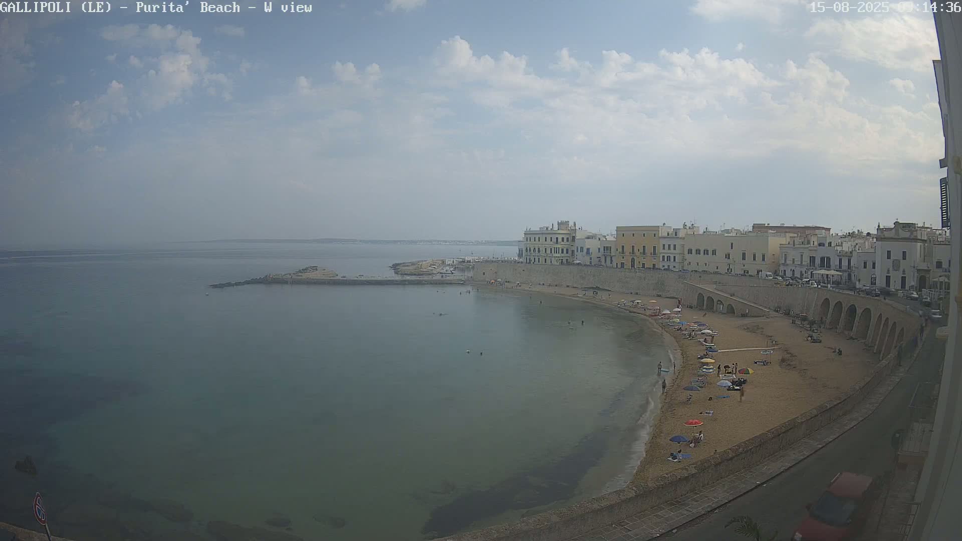 A partly cloudy day shows a sandy beach curving around a calm bay, lined with low buildings and people enjoying the sun.