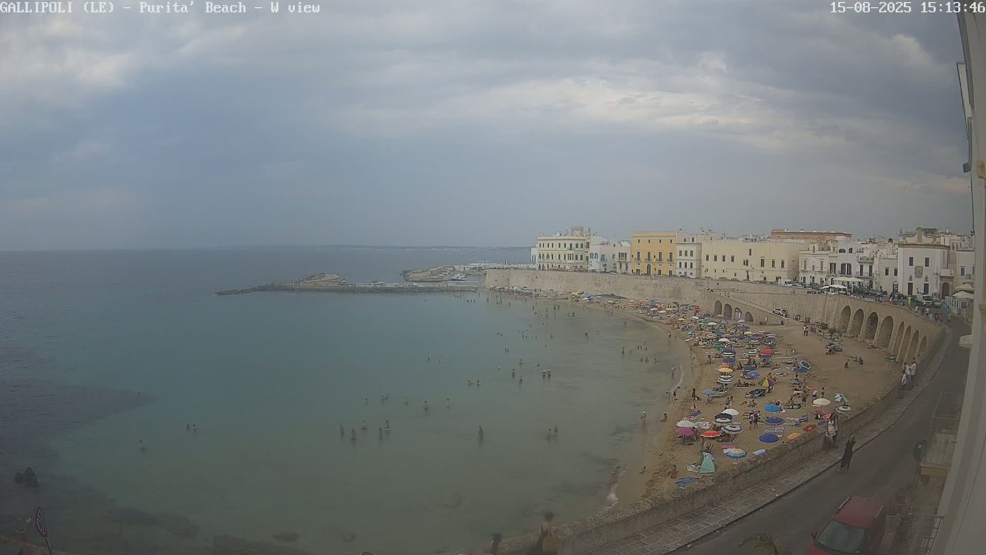 A partly cloudy day shows a sandy beach curving around a calm bay, lined with low buildings and people enjoying the sun.
