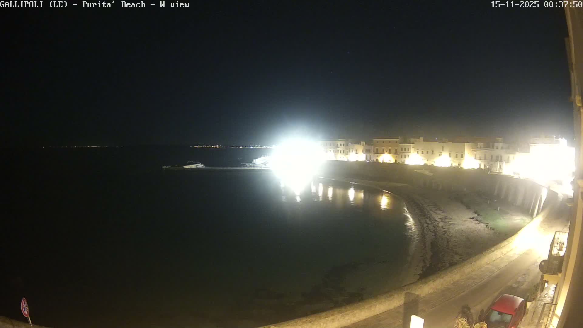 On a clear, calm night, a coastal scene reveals illuminated buildings and a sandy beach lining calm waters, where a blinding light source creates a prominent reflection, with a boat visible offshore.