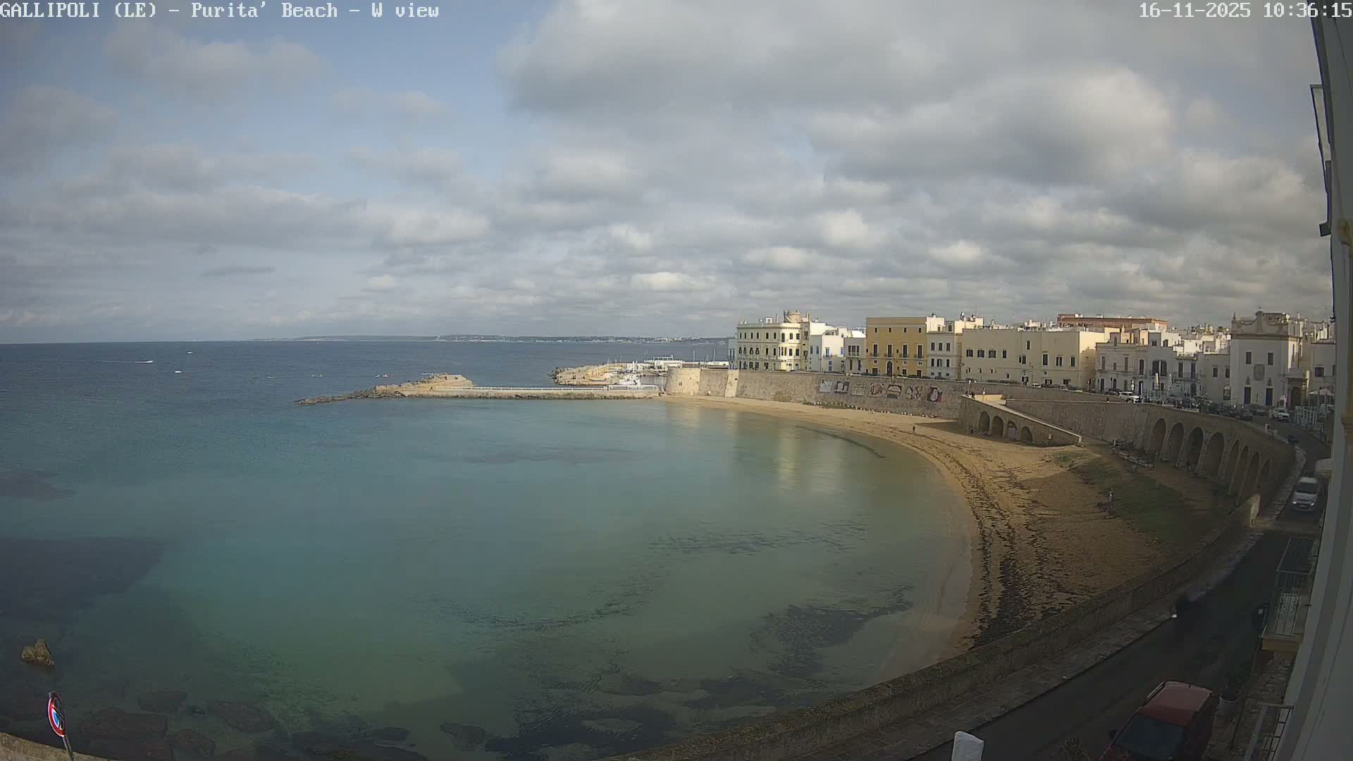 A picturesque coastal town featuring a curved sandy beach, clear blue-green water, and numerous light-colored buildings along a stone promenade is captured under a partly cloudy sky.