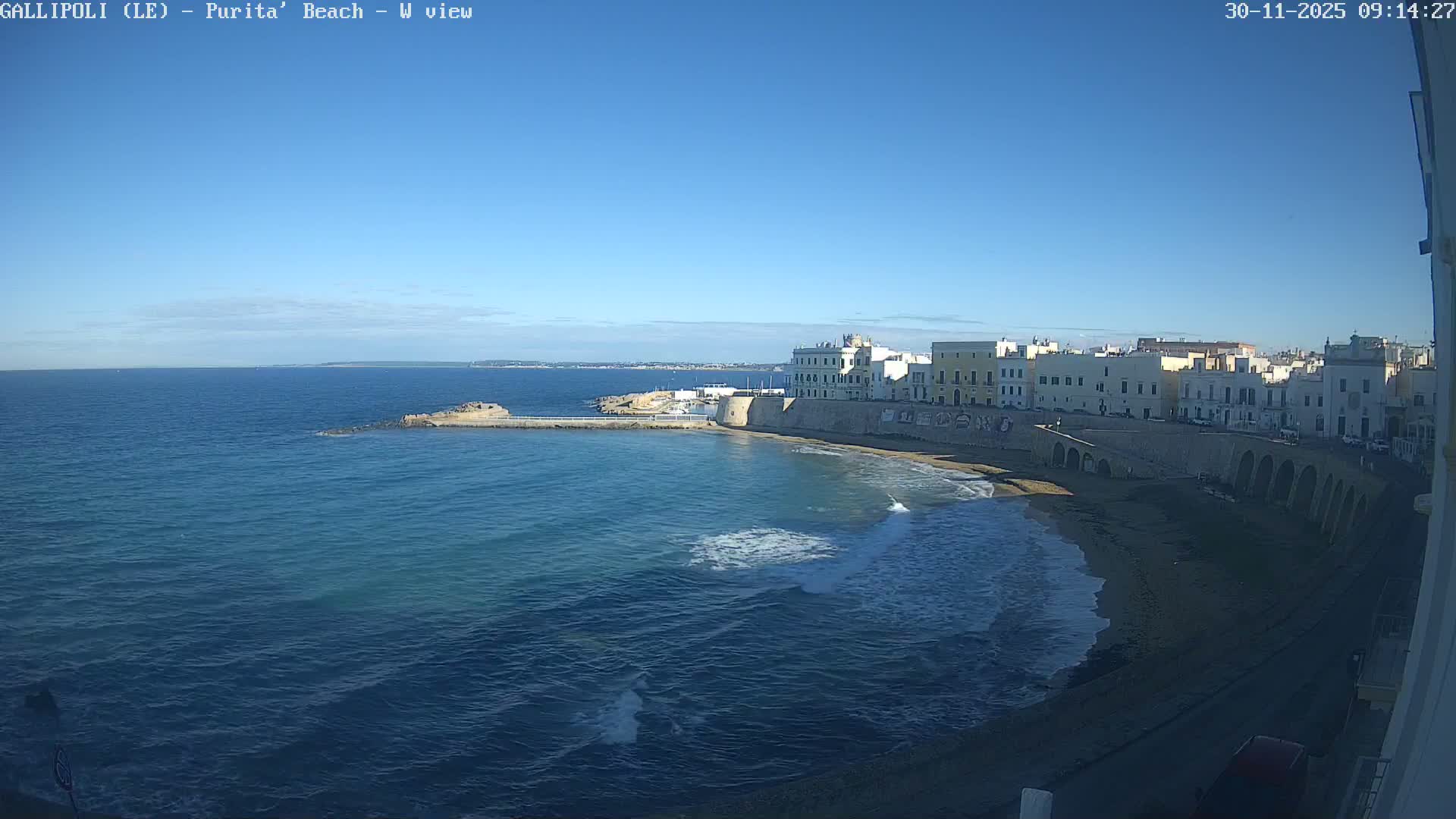 Overlooking a coastal town on a bright, sunny day, the image features a curved beach with turquoise waves breaking against a stone seawall and a small pier, backed by white buildings, with distant land visible across the calm blue sea.
