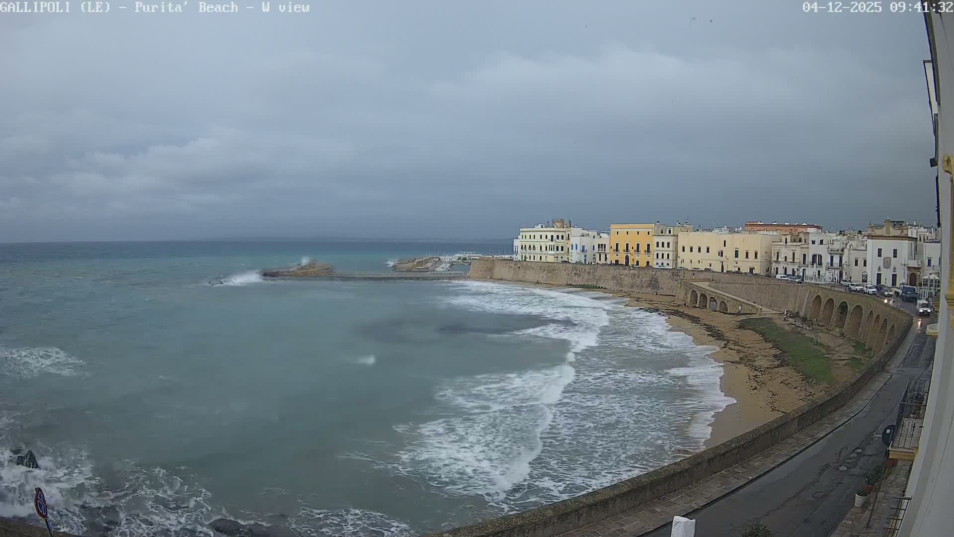 Large waves crash against the narrow beach and a distant pier of a coastal town lined with historic buildings and a wet road, all under a dark, overcast sky.