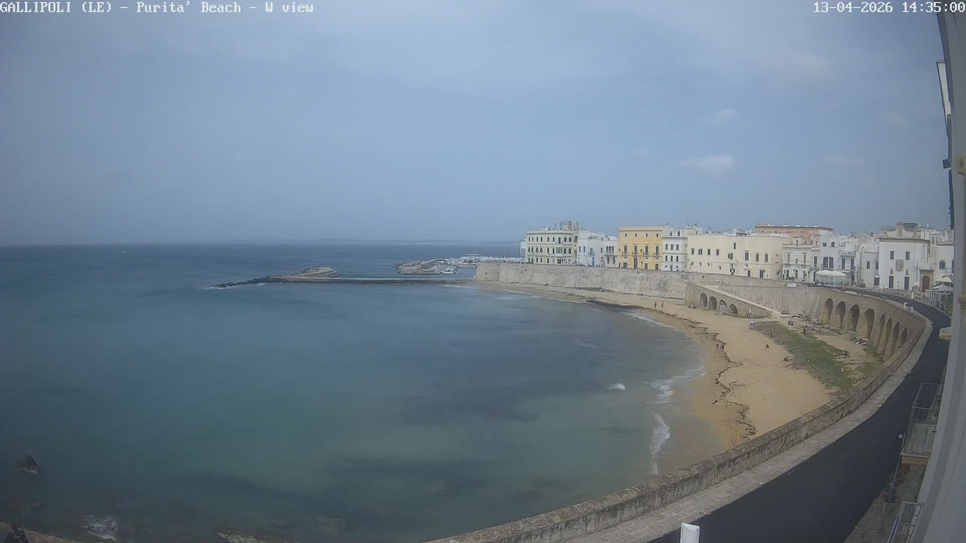 Large waves crash against the narrow beach and a distant pier of a coastal town lined with historic buildings and a wet road, all under a dark, overcast sky.