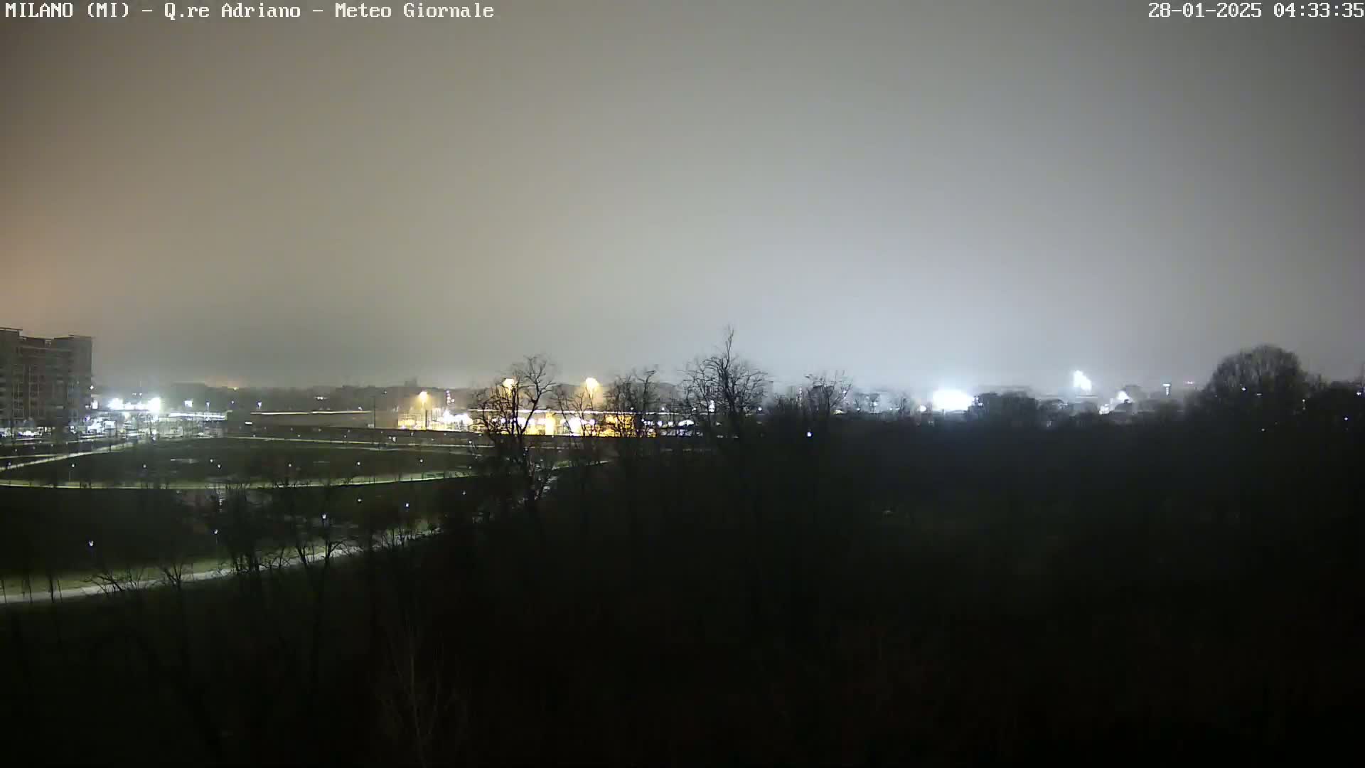 A nighttime, hazy view of a city park and buildings, dimly lit with artificial lights.