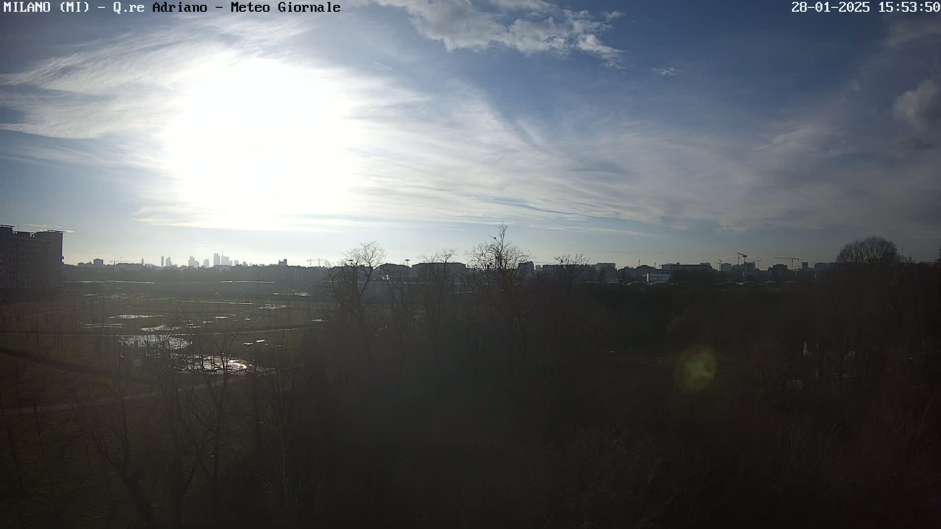 A mostly sunny day reveals a cityscape in the distance, partially obscured by a dark foreground of trees and a flat, somewhat marshy area.