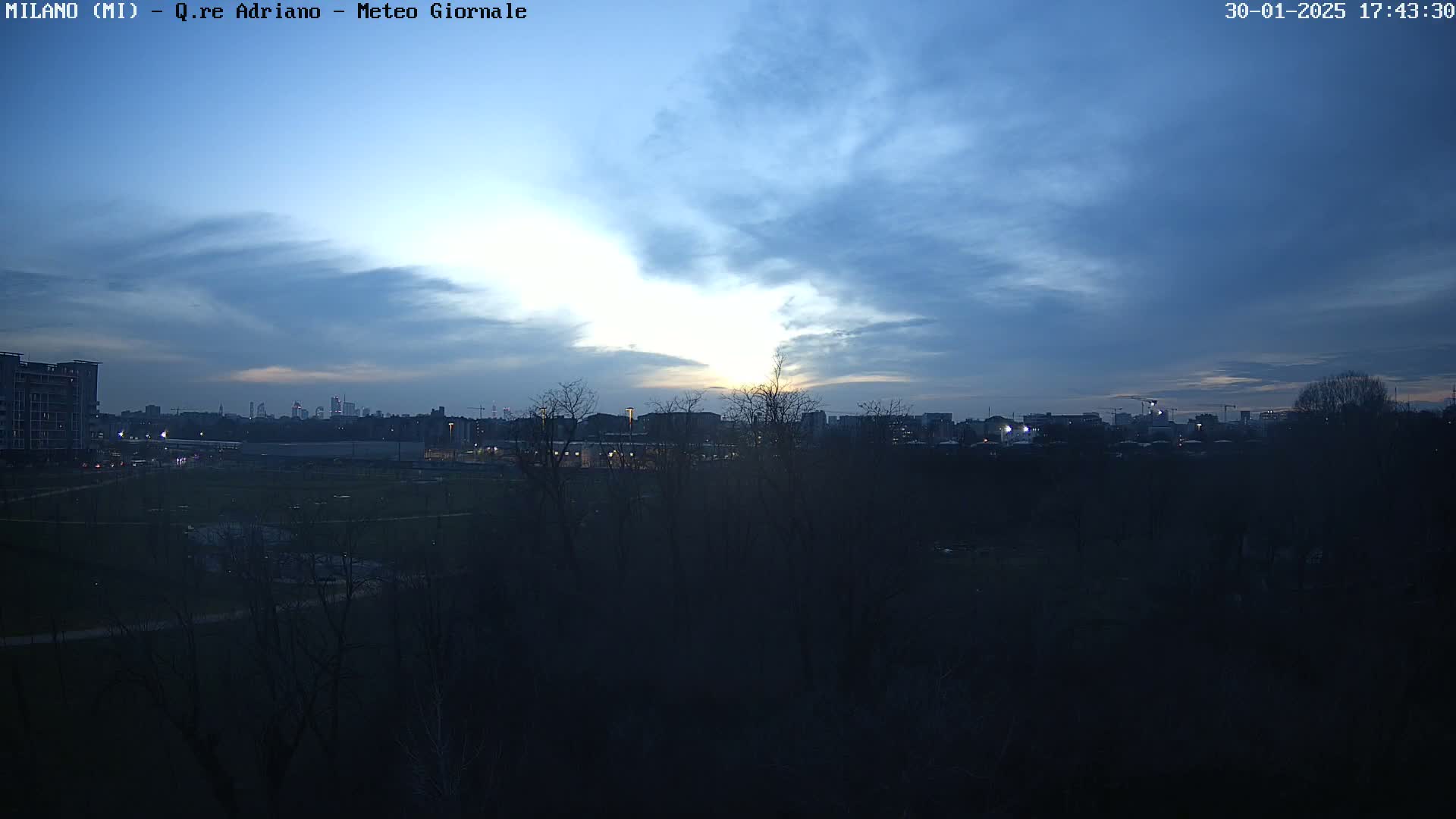 A mostly cloudy, twilight sky over a city skyline viewed from across a dark, leafless park.
