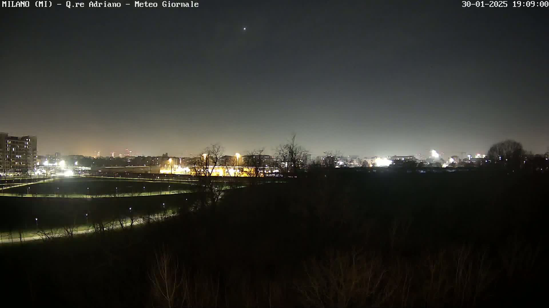 A nighttime view of a city skyline across a dark, sparsely vegetated area under a clear, dark sky.