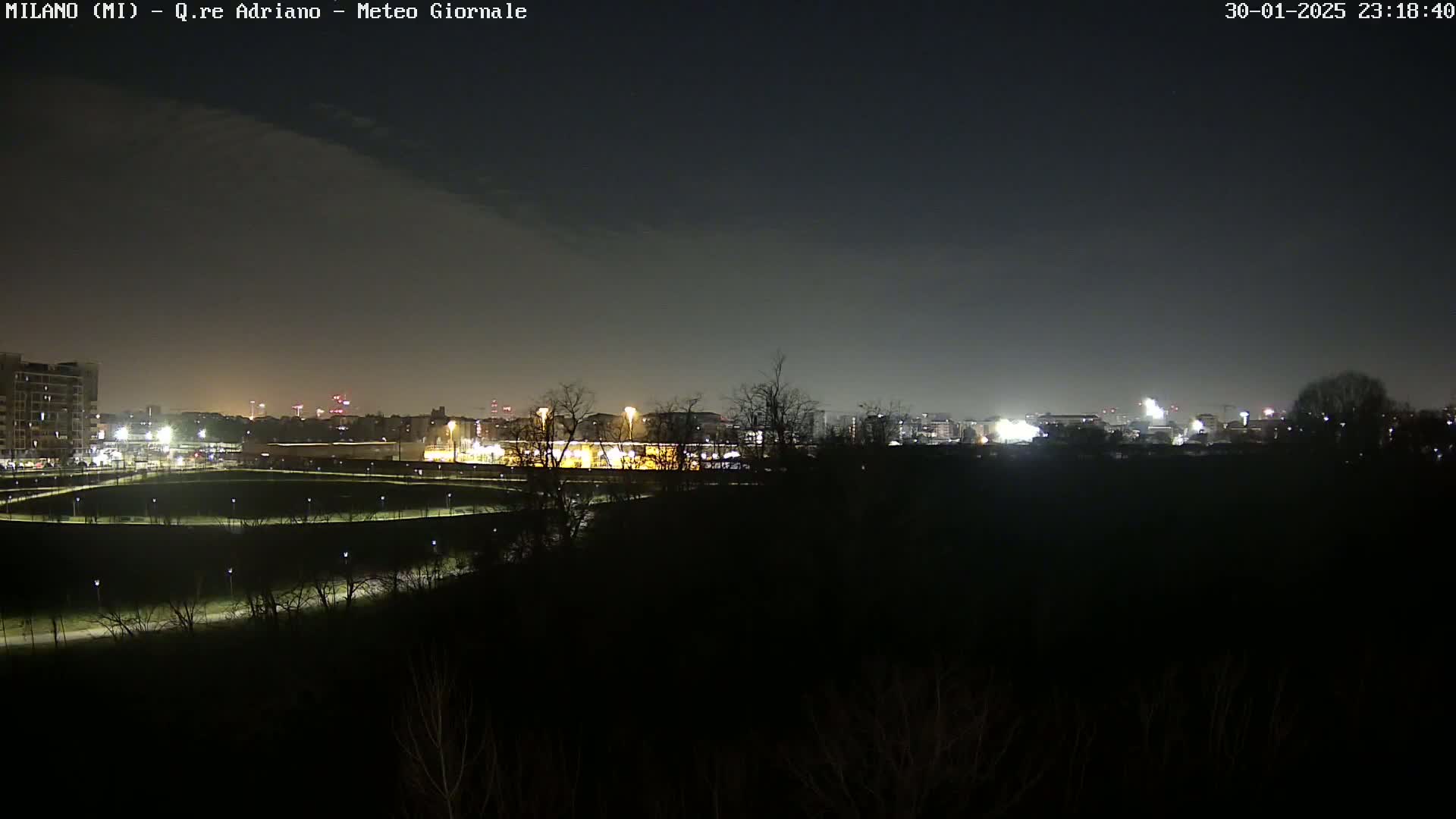 A nighttime view of a city skyline across a dark, treeline landscape under a clear, dark sky.