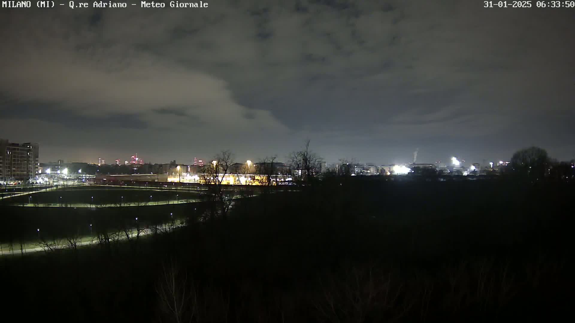 A nighttime view of a city skyline across a dark, tree-lined park under a cloudy sky.