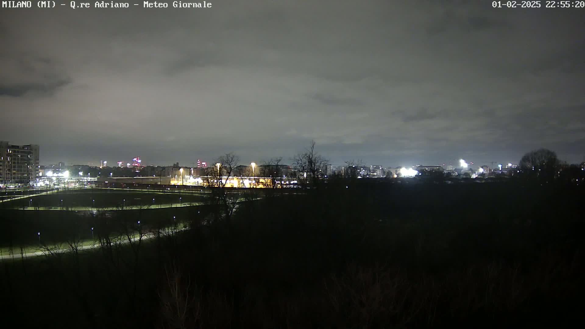 A nighttime view of a city skyline across a dark, tree-covered landscape under a cloudy sky.