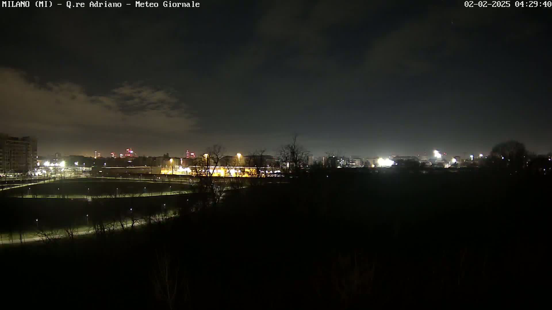 A nighttime view of a city skyline across a dark, sparsely lit park under a mostly cloudy sky.