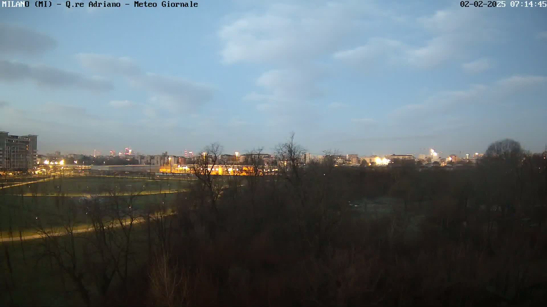 A city skyline is visible at dusk under a mostly clear sky, with a park and trees in the foreground.