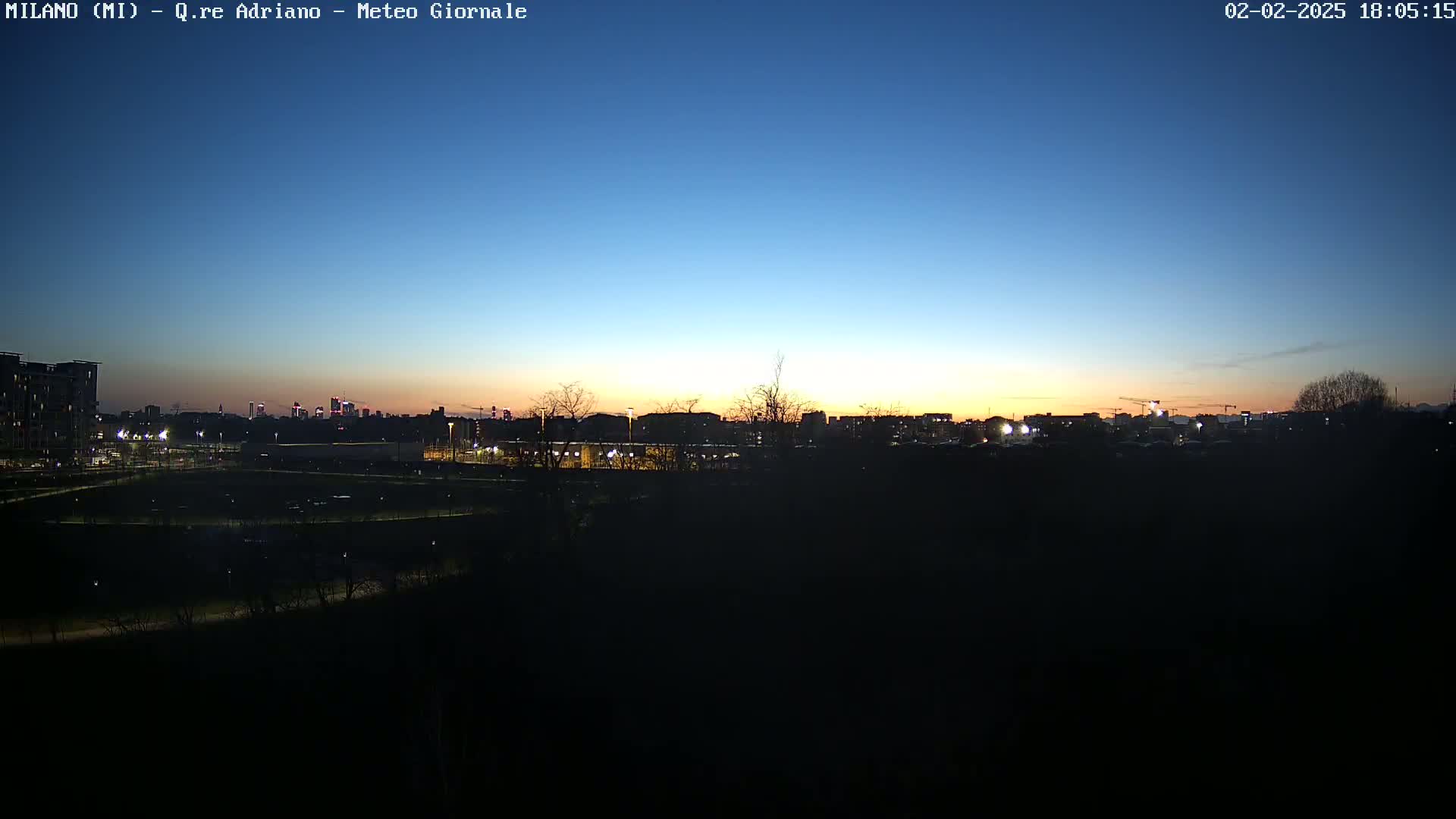 A city skyline is visible at sunset under a clear twilight sky, viewed from a dark, sparsely vegetated area.