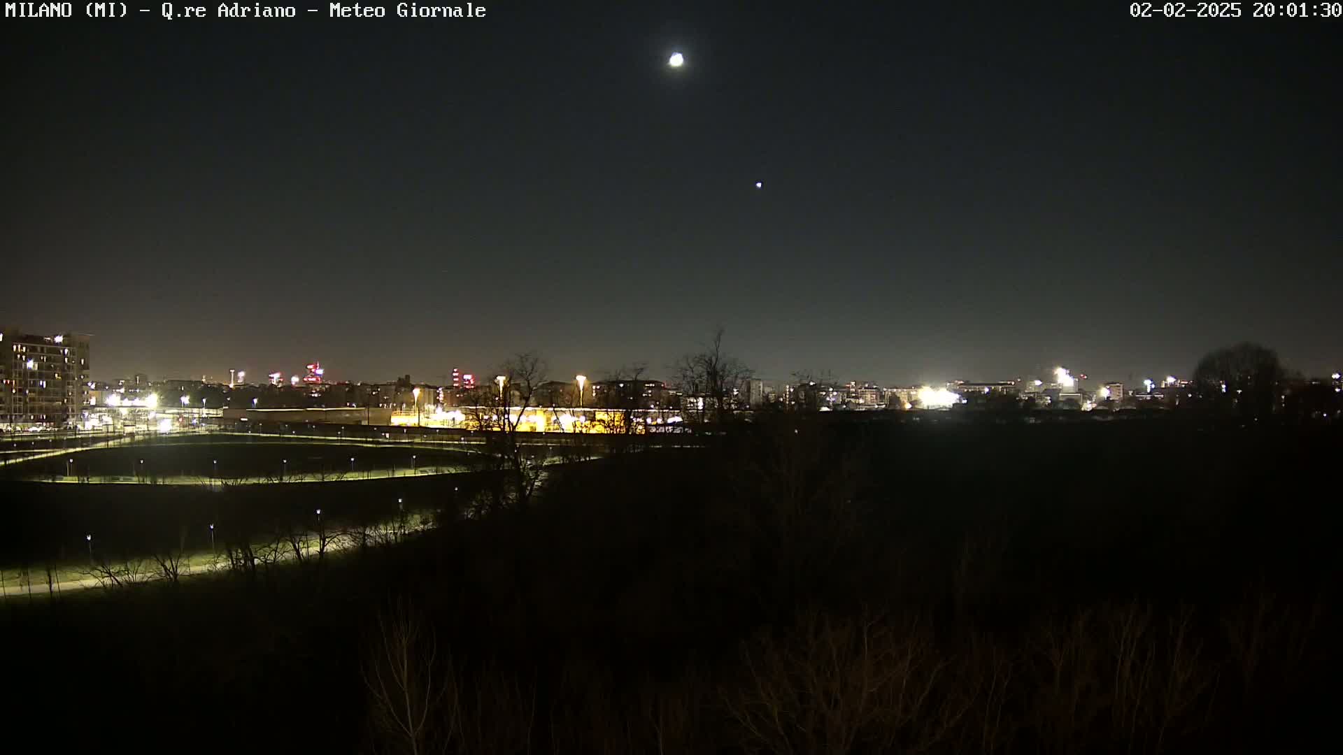 A nighttime view of a city skyline illuminated by lights, with a moon and a star visible in a clear night sky.