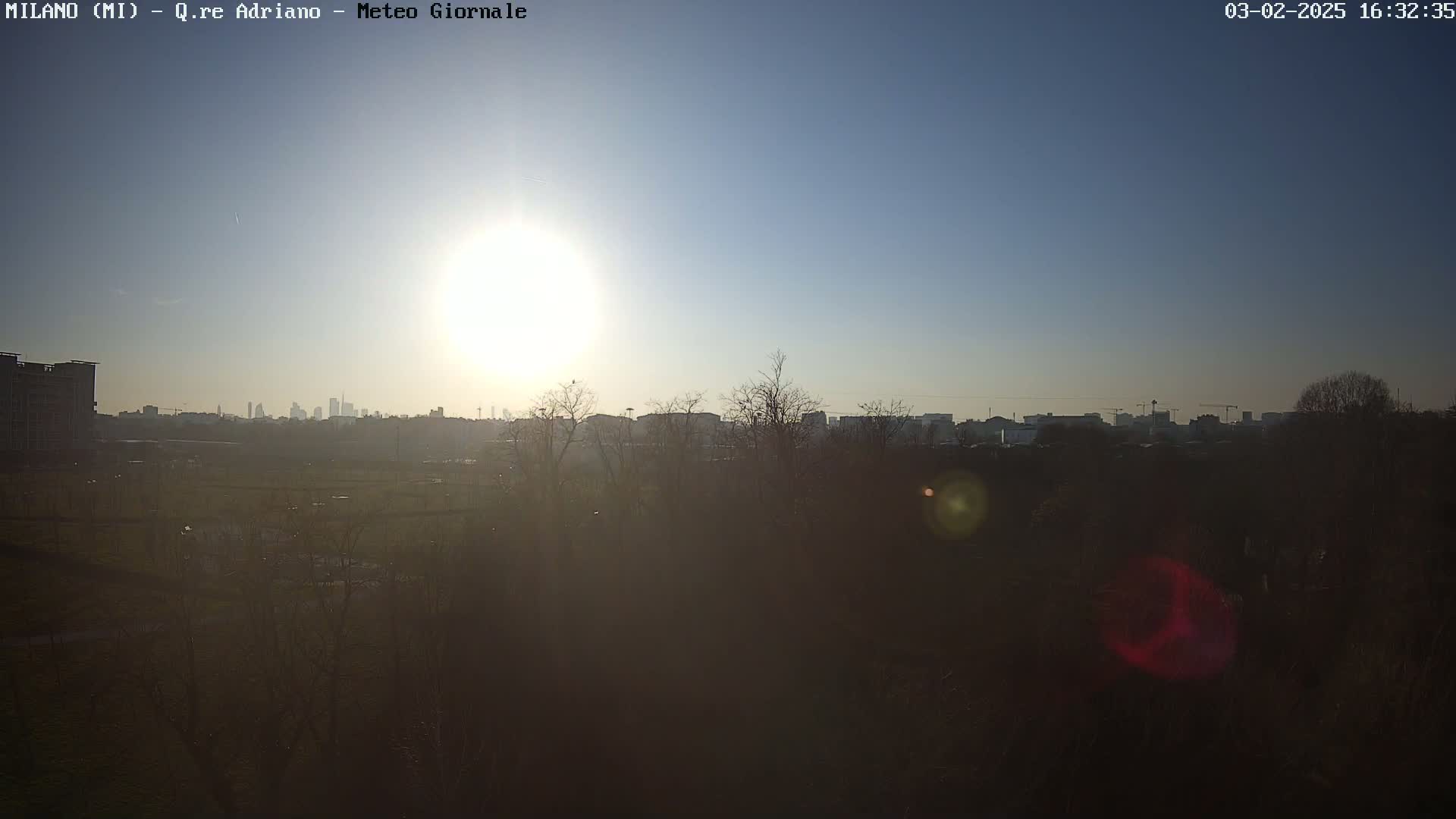 A bright sun shines in a clear blue sky over a city skyline partially obscured by dark, leafless trees and a field in the foreground.