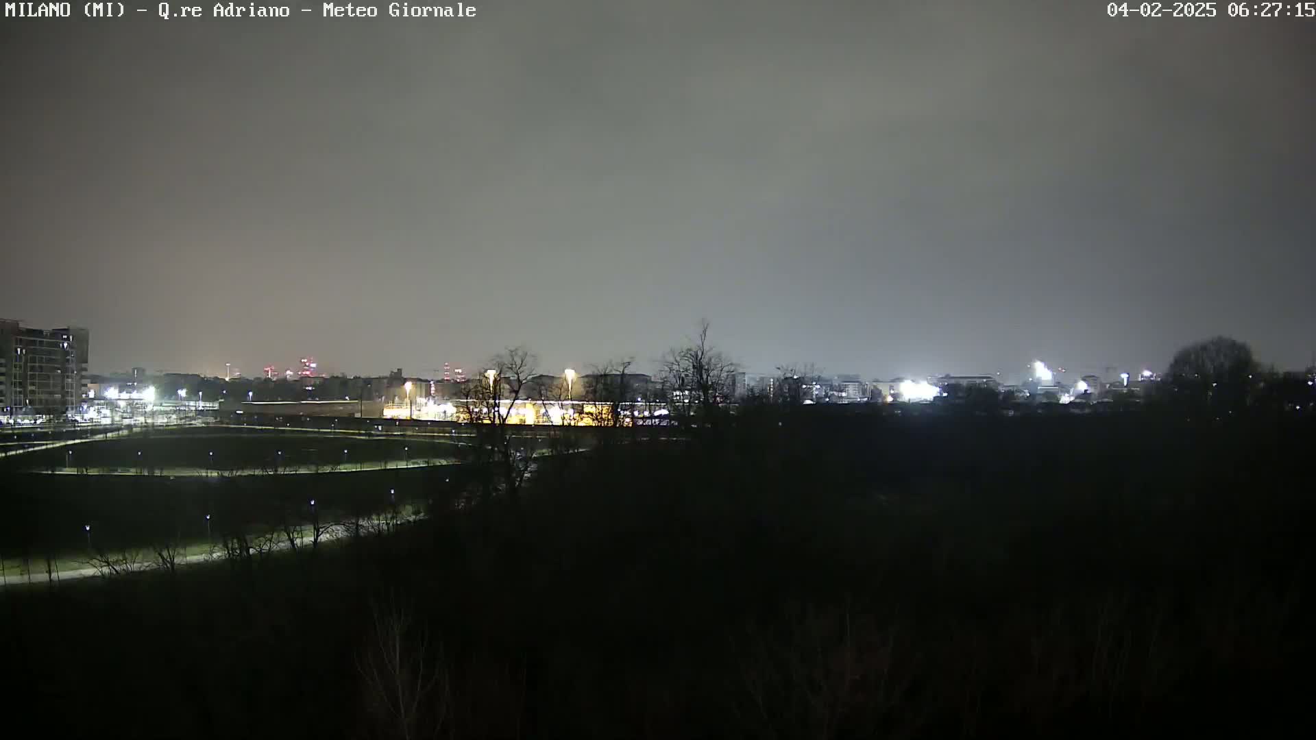 A nighttime view of a city skyline under an overcast sky, showing dimly lit buildings and trees in the foreground.