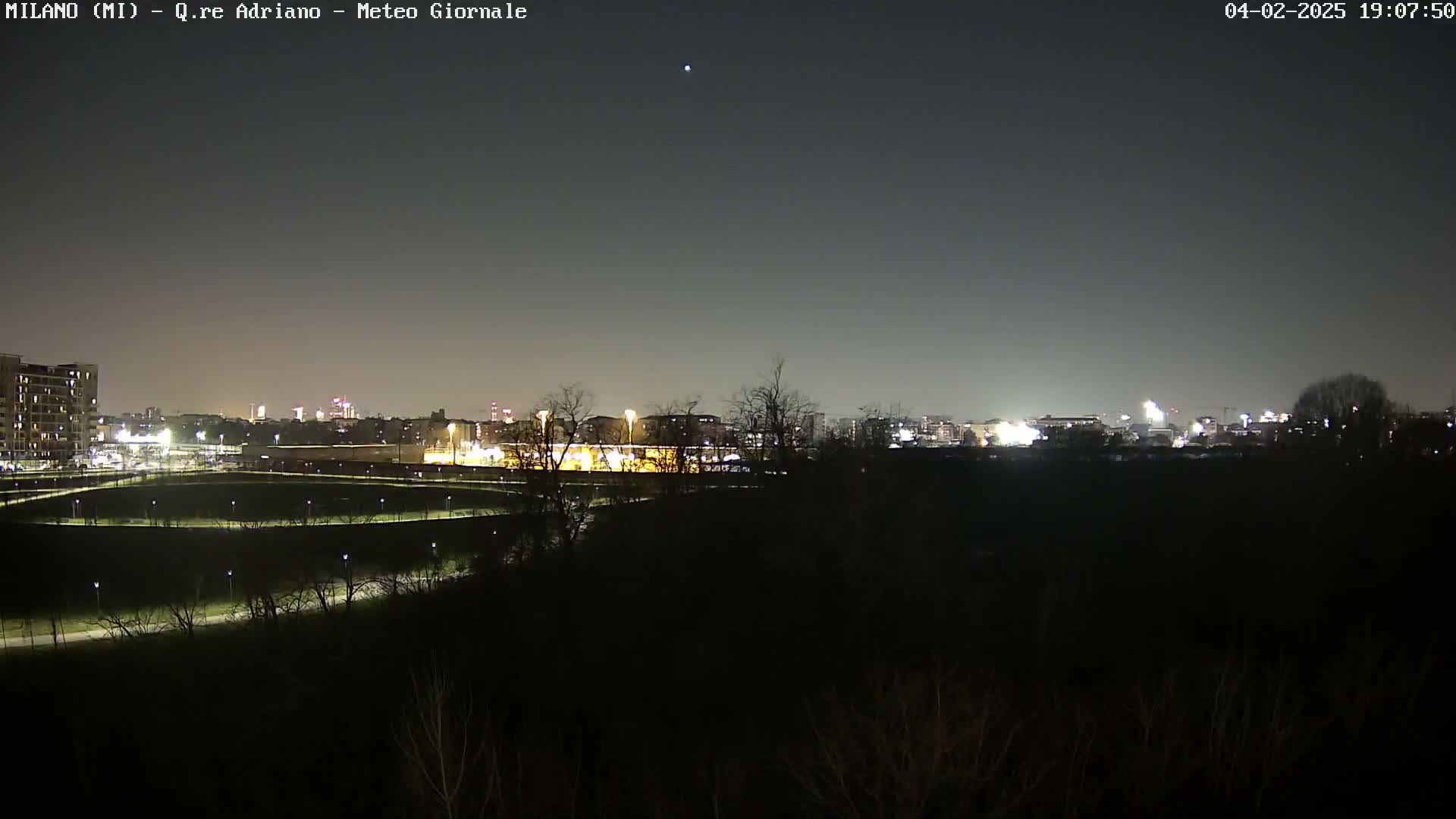 A nighttime view of a city skyline across a dark, sparsely lit park, under a clear, dark sky.