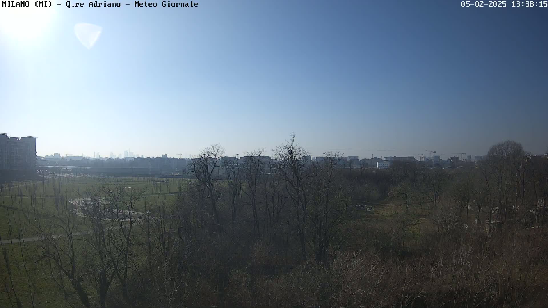 A mostly clear blue sky overlooks a flat, grassy field dotted with bare winter trees, beyond which lies a cityscape in the distance.