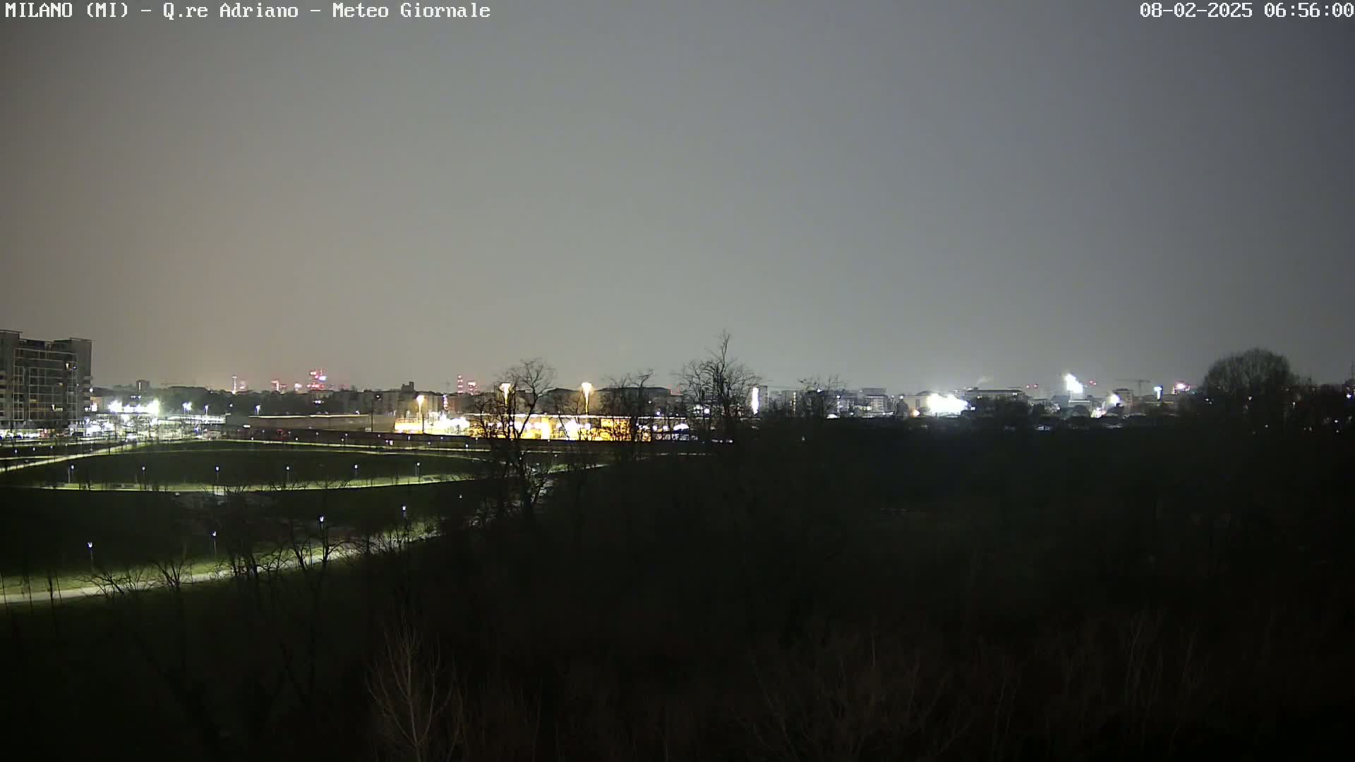 A nighttime view of a city skyline dimly lit, seen from across a dark, sparsely lit park under a gray, overcast sky.