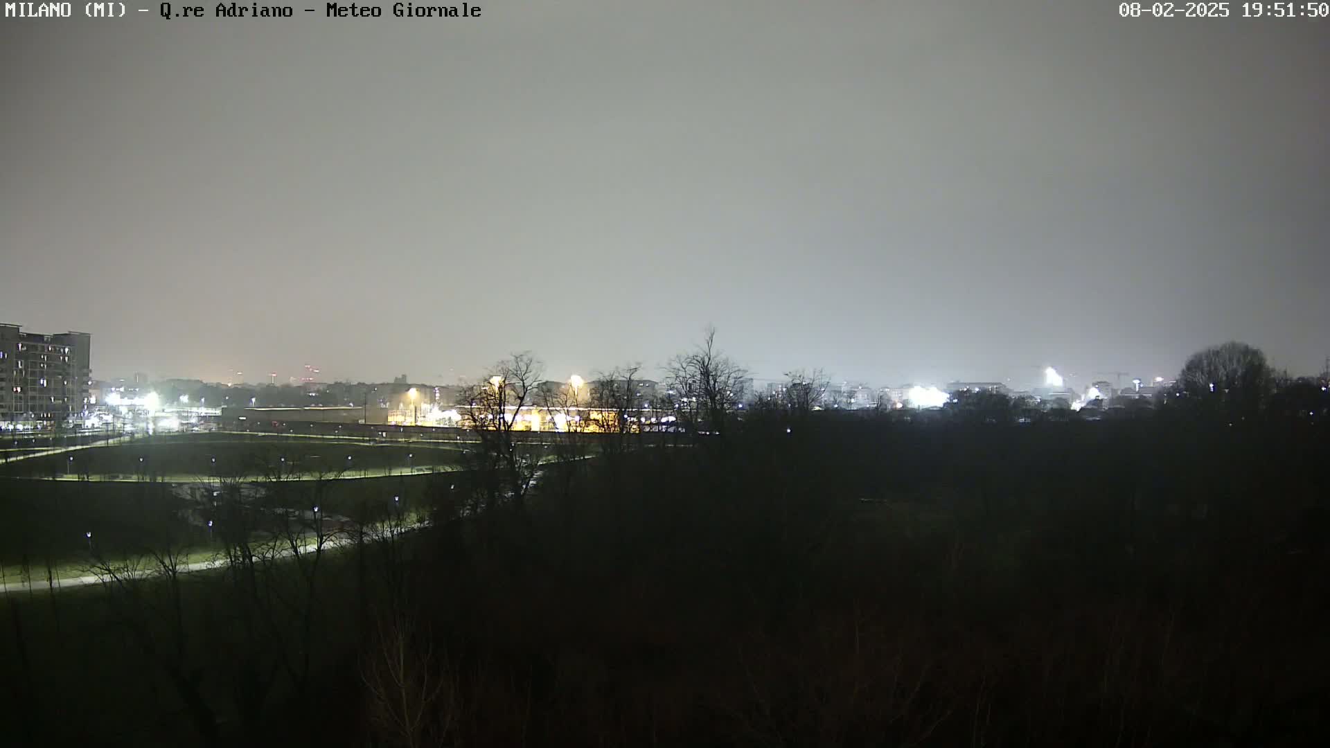 A nighttime view of a city skyline dimly lit under a hazy, overcast sky, seen from across a dark park with sparsely lit pathways.