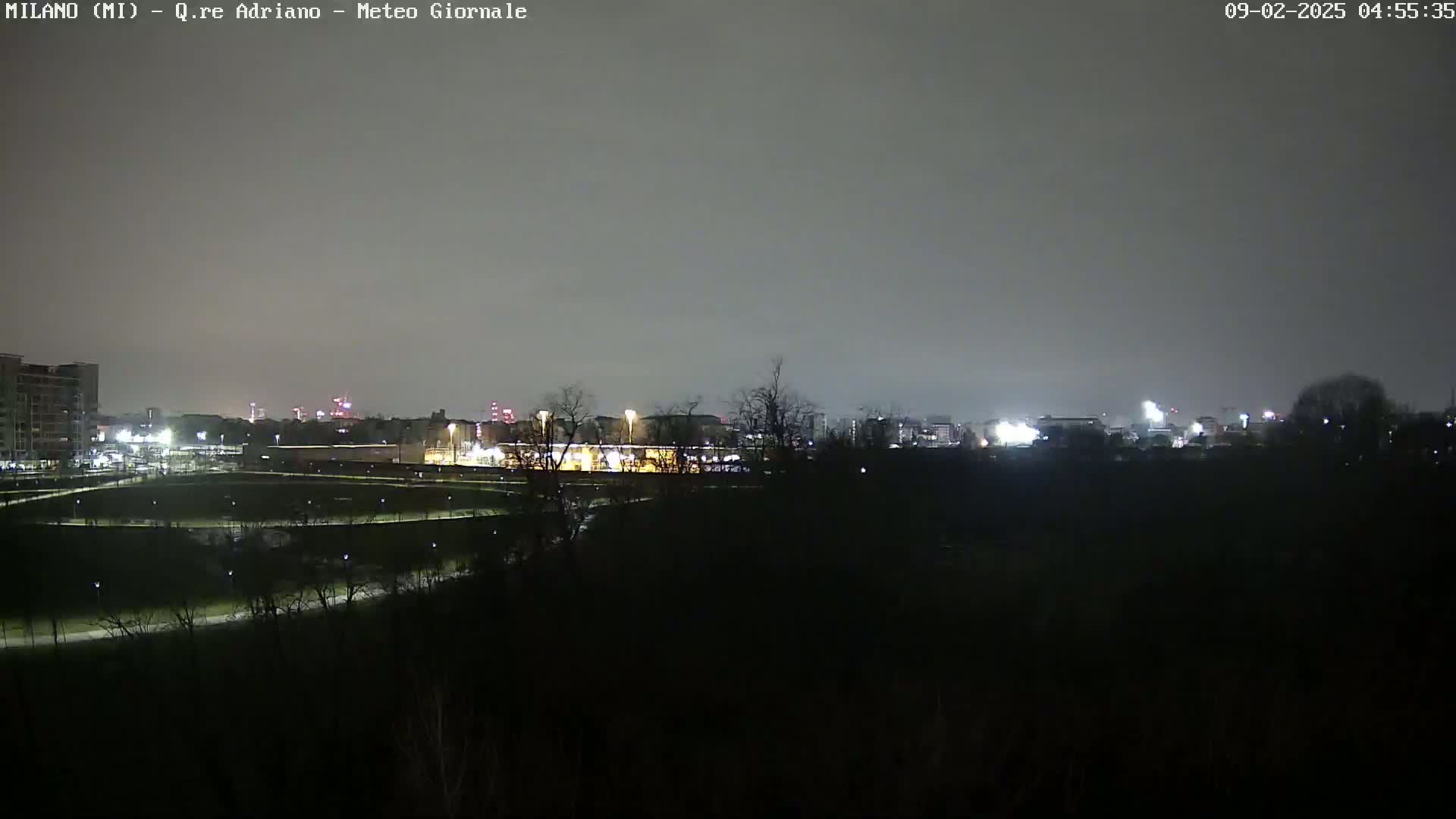 A nighttime view of a city skyline under a cloudy sky, seen from across a dark expanse of land with some lights illuminating paths.