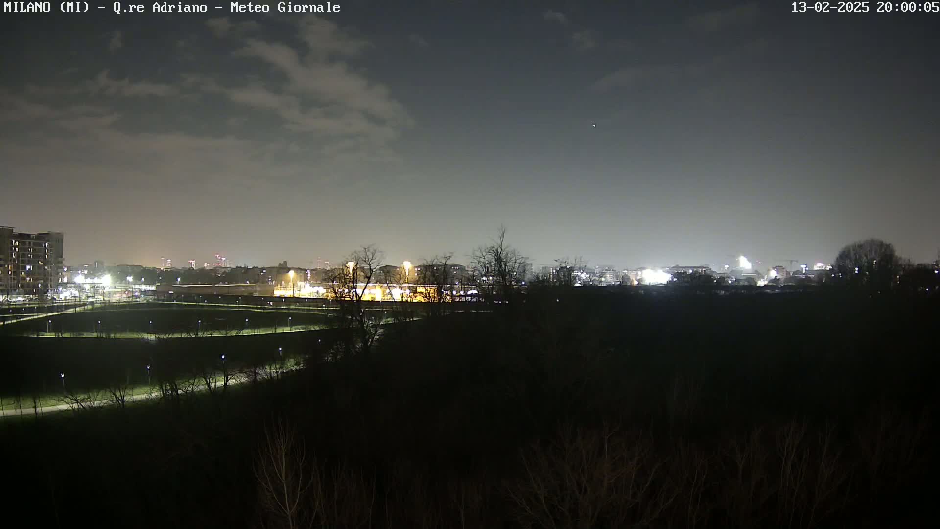 A nighttime view of a city skyline across a dark, sparsely lit park under a cloudy sky.