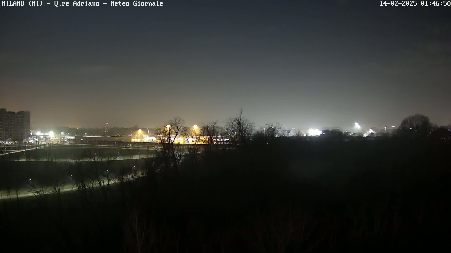 A nighttime, hazy view of a park with dimly lit paths, buildings, and trees in the distance under a dark sky.