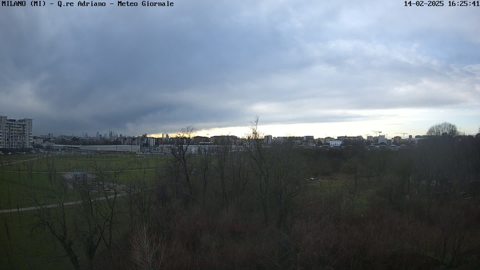 A mostly cloudy sky hangs over a park with leafless trees and buildings in the distance.