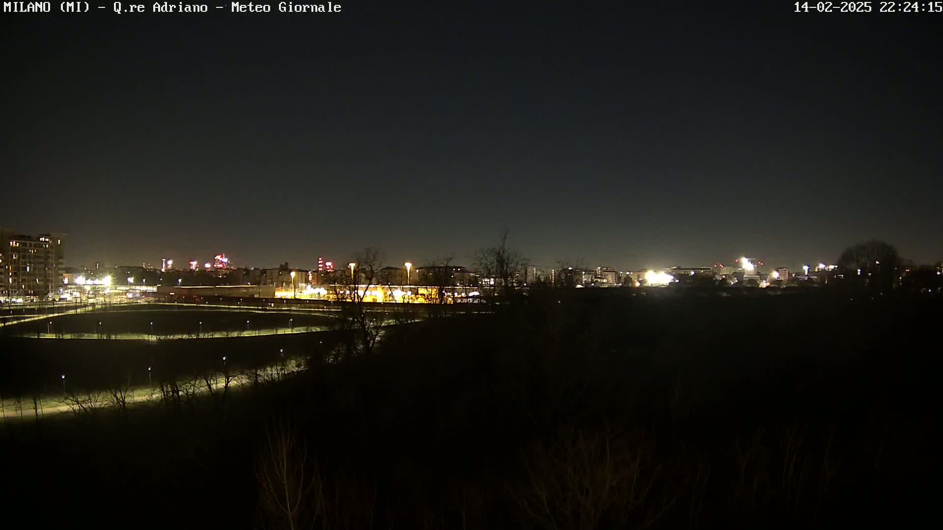 A nighttime view of a city skyline across a dark field, with dimly lit trees and paths in the foreground under a clear, dark sky.