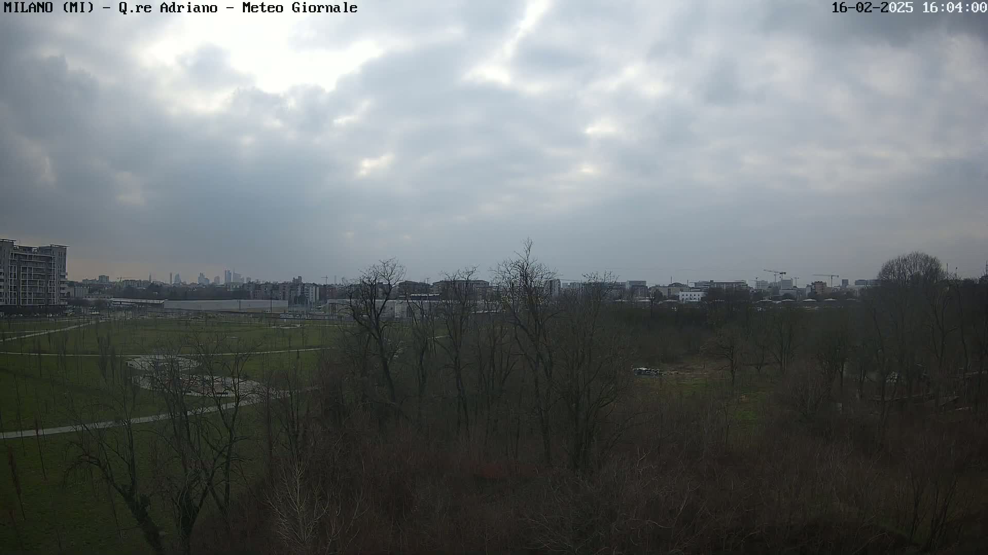A mostly cloudy sky overlooks a park with bare trees and buildings in the distance.