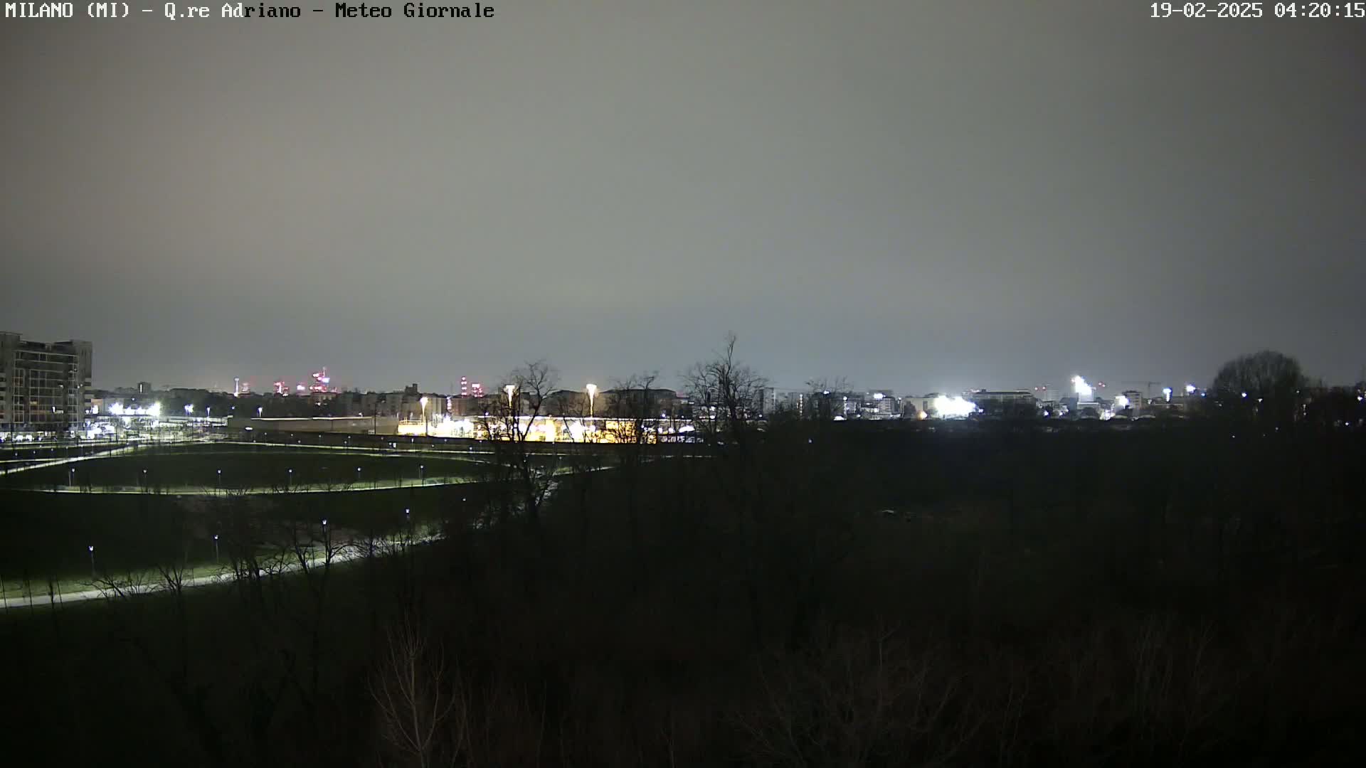 A nighttime view of a park with dimly lit pathways and trees in the foreground, overlooking a city skyline under a cloudy sky.