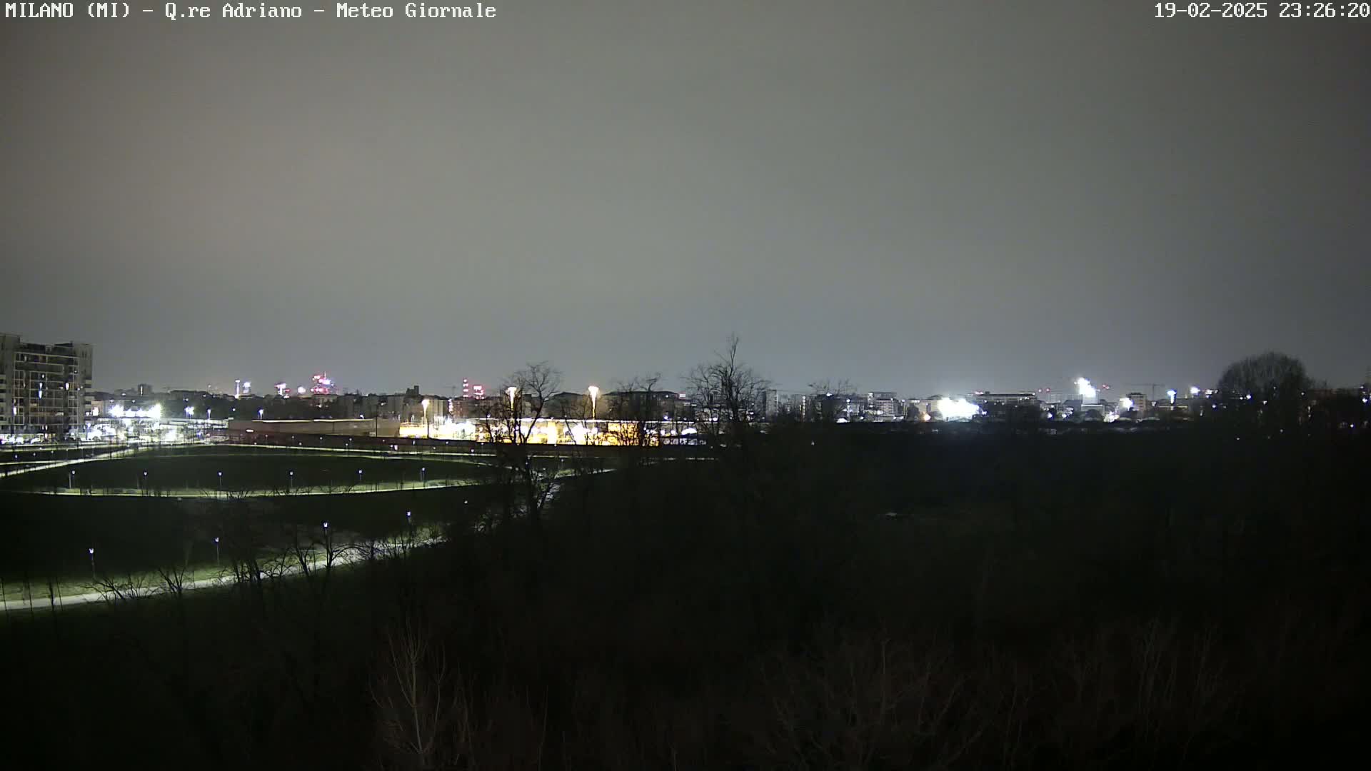 A nighttime view of a city skyline across a dark, sparsely lit park under a cloudy sky.