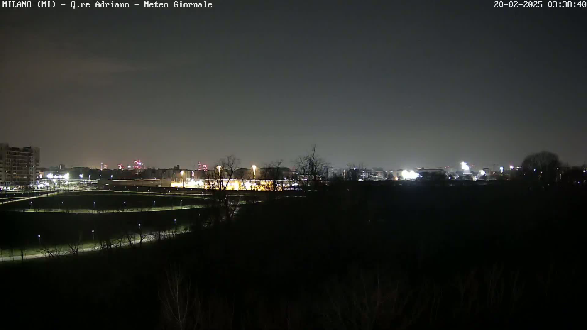 A nighttime view of a city skyline across a dark, flat expanse of land with sparsely lit paths and trees.