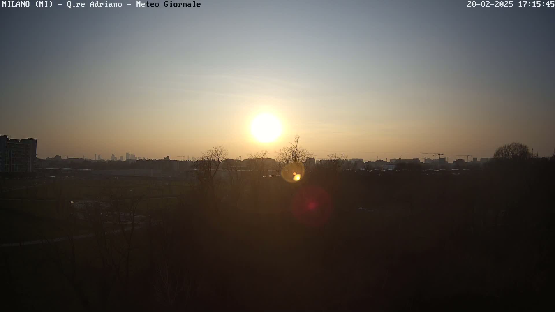 A sunset over a city skyline, partially obscured by bare trees and a hazy atmosphere.