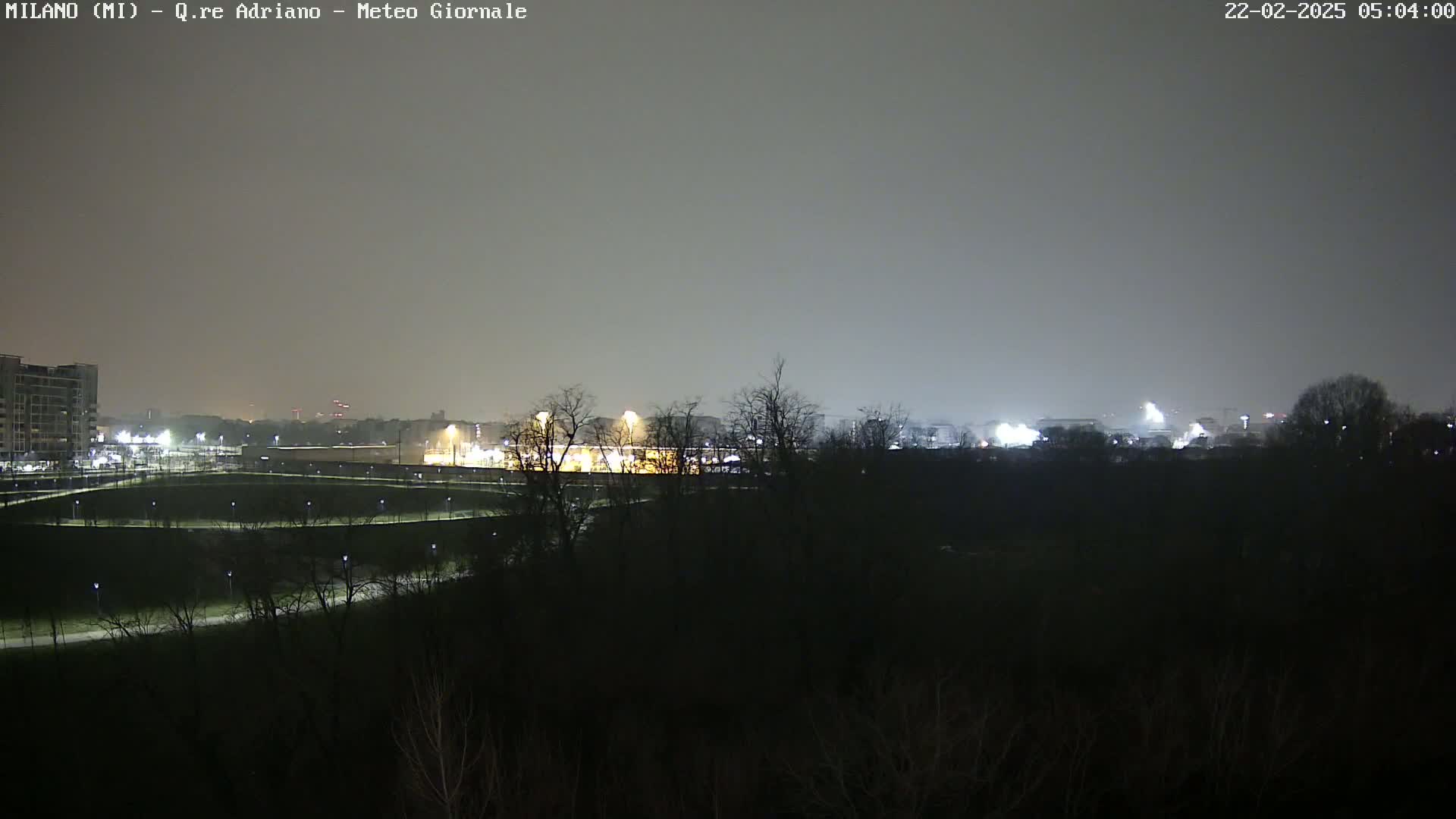 A nighttime view of a park with dimly lit pathways and buildings in the distance under a hazy sky.