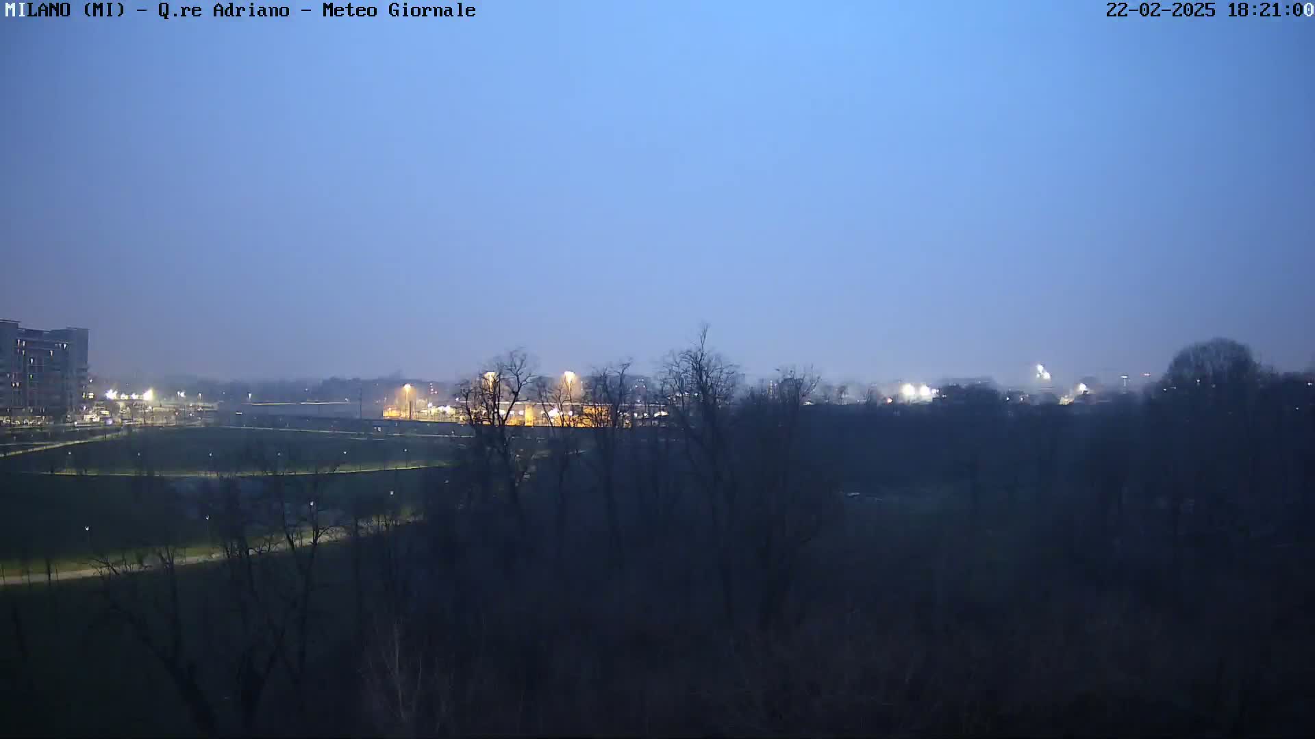 A twilight view shows a flat expanse of land with bare trees, sparsely lit buildings in the distance, and a clear, dark blue sky.