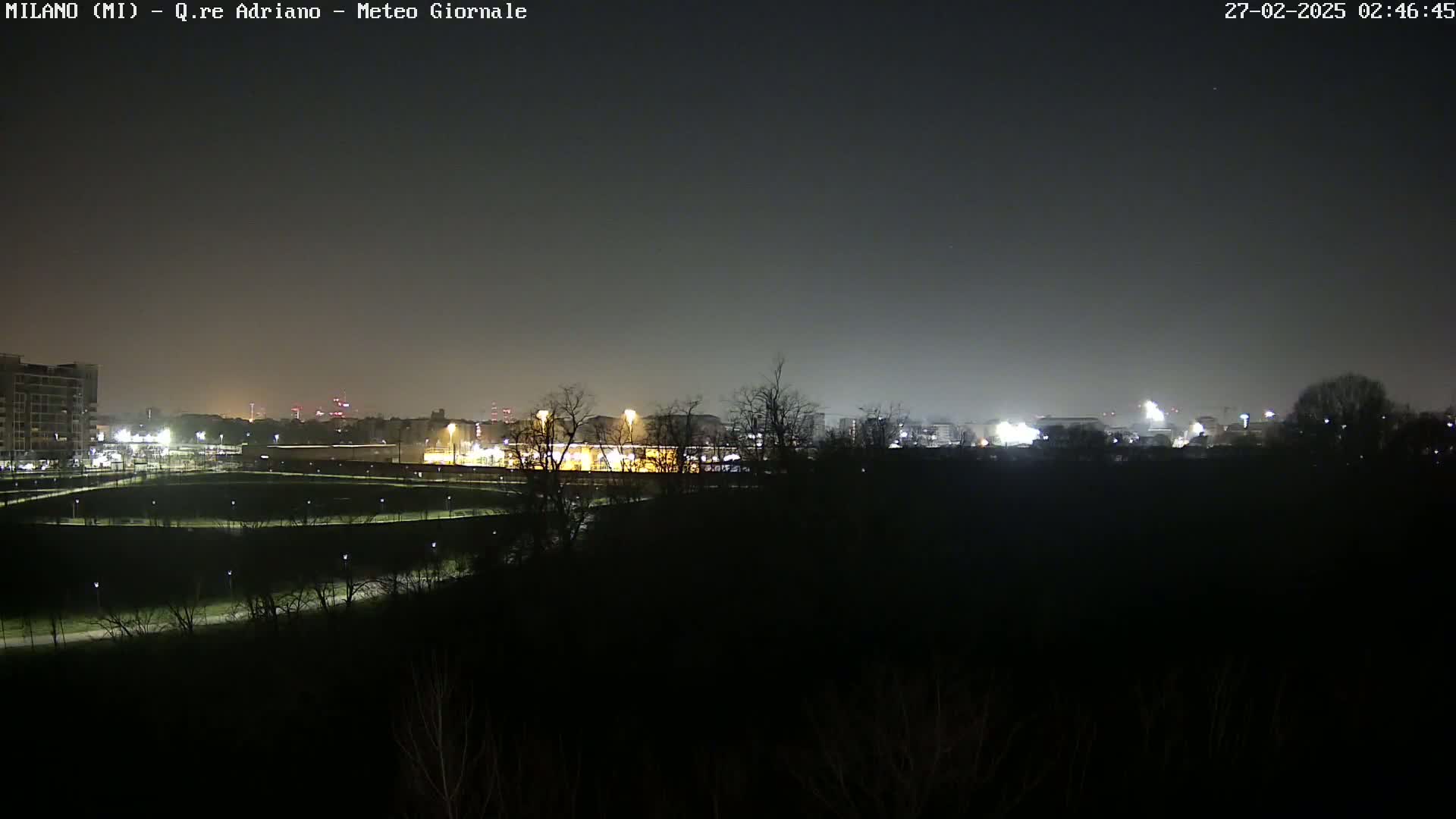 A nighttime view of a city skyline dimly lit across a dark expanse of land with bare trees under a dark sky.